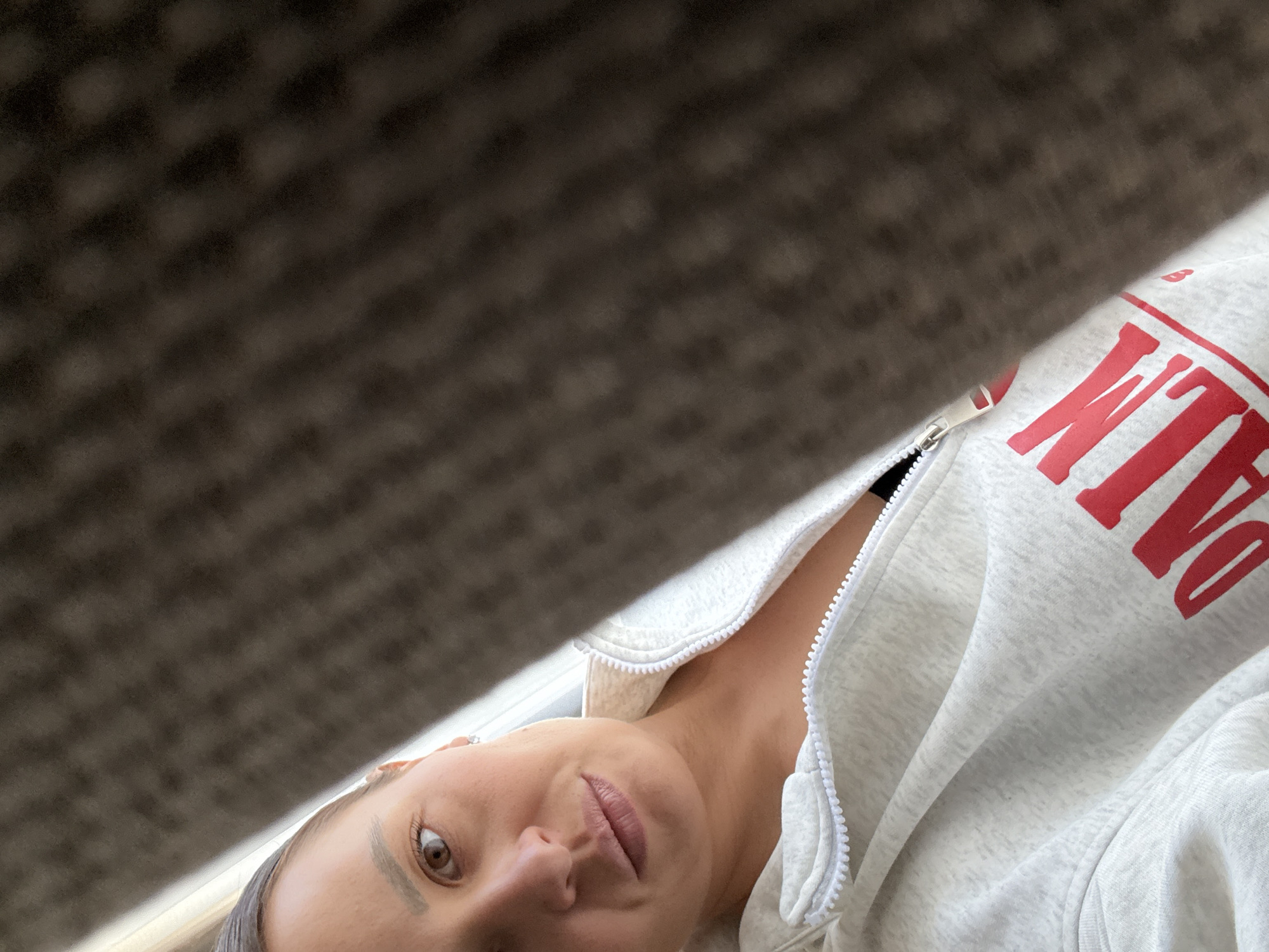 A woman is looking down at the camera, partially visible in the lower portion of the frame. She is wearing a gray hooded sweatshirt with a white zipper and red lettering. The top portion of the frame is filled with a blurred, textured brown surface. The location is not discernible from the visual information provided.