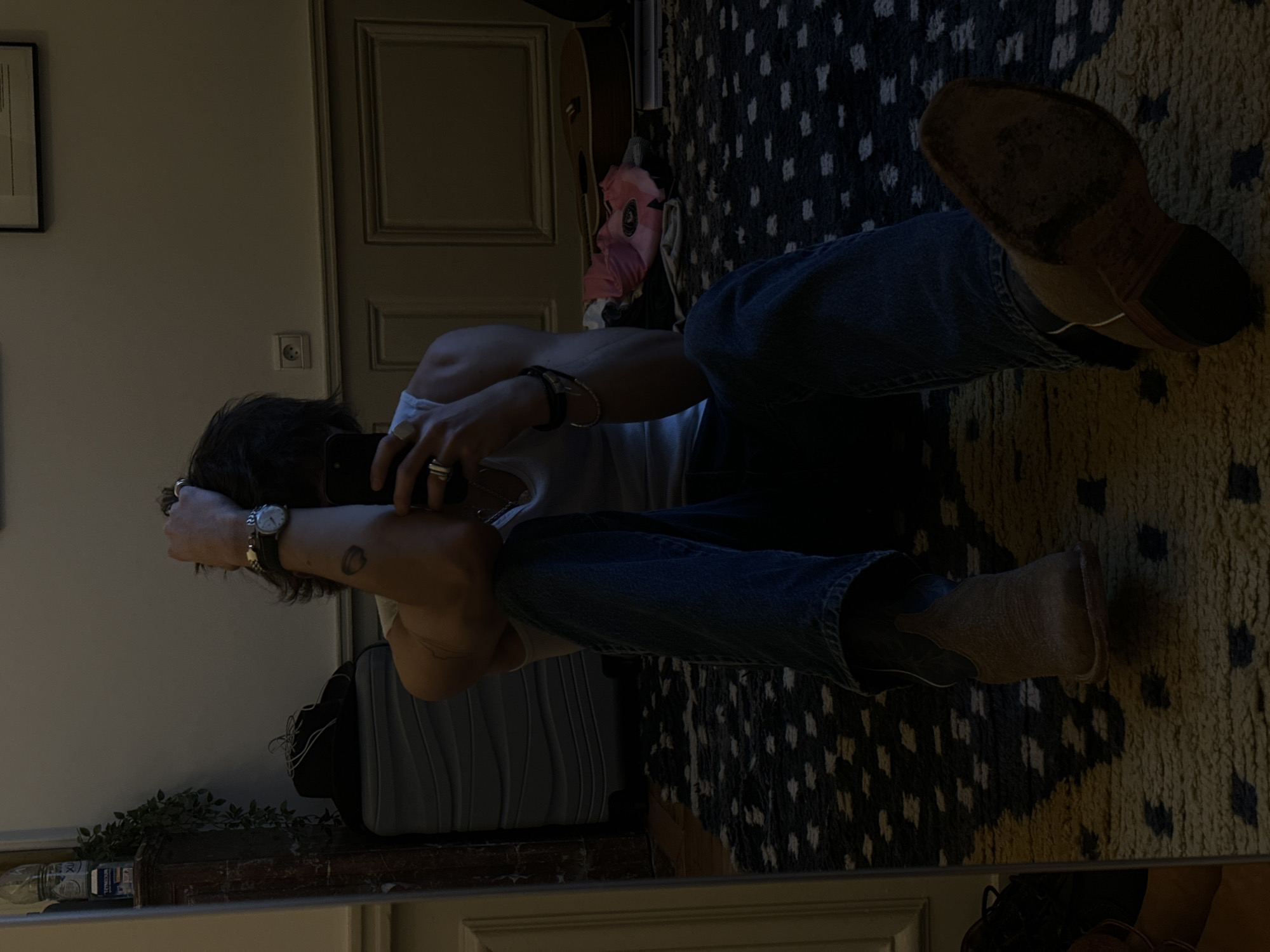 A person is seated on a patterned rug in a room with classic Parisian architectural details, including paneled doors and ornate molding. The person is wearing jeans, a white tank top, and cowboy boots, and is looking at their phone. There is a guitar leaning against the wall, and a suitcase is nearby. The room also contains a decorative mantelpiece with plants.