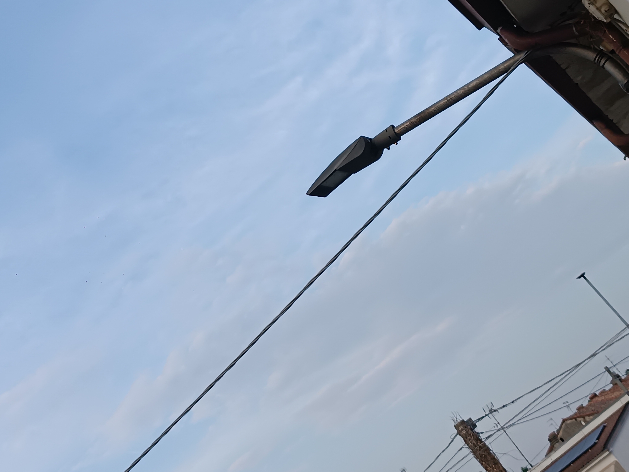 The image captures a slightly tilted, close-up view of a streetlamp angled against a pale blue sky with wispy clouds. The streetlamp is dark and sleek, mounted on a metallic pole that extends towards the top right corner of the frame. Below and to the right, a cluster of power lines crisscrosses the sky, with a distant view of rooftops and a smaller, thinner streetlamp further in the distance. The scene appears to be taken outdoors during the day, possibly in the city of Viadana, Italy, given the architectural style of the visible rooftops. There are no people or immediate action depicted, creating a static and somewhat impersonal urban landscape.