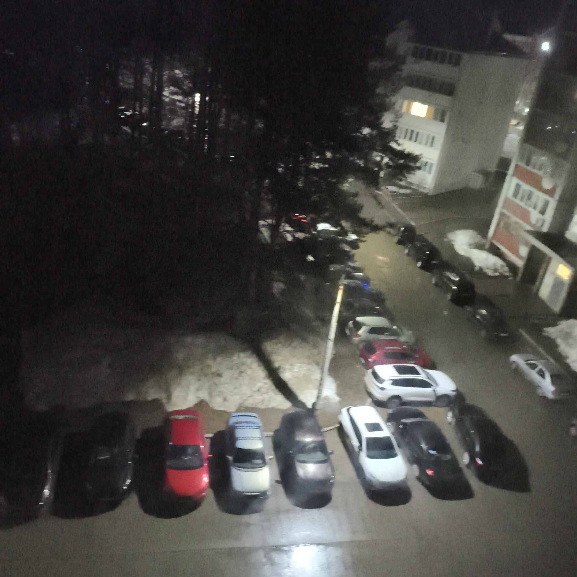 The image shows a night scene with a parking lot filled with cars. In the background, there are buildings, trees, and snow patches. The lighting is dim, primarily from streetlights and building lights, creating strong shadows from the cars and trees. The parking lot contains a variety of cars, distinguished by color and size. The general location appears to be a residential or commercial area with a parking facility.