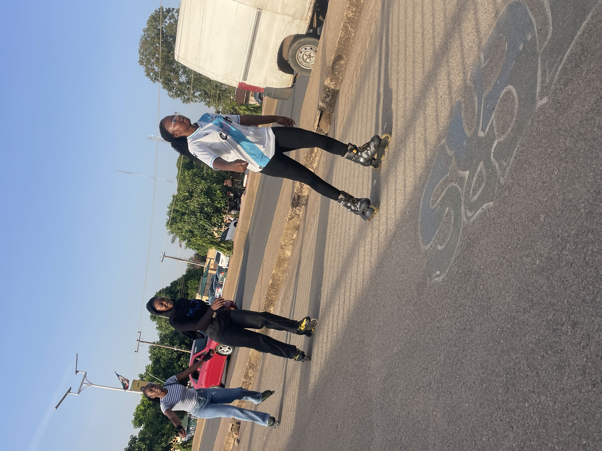 The image depicts three individuals on a paved road or street under a clear, bright blue sky. The lighting suggests either early morning or late afternoon, as long shadows are cast across the asphalt.
In the foreground, a young woman with dark braided hair and glasses is seen. She wears a white and light blue striped jersey, characteristic of the Argentina national football team, paired with black leggings and black inline skates. She stands balanced on her skates, looking upwards with a relaxed expression.
Further back and to the left, another person is dressed in a black long-sleeved top, black trousers, and black inline skates with yellow accents, appearing to be in motion. Behind them and slightly to the left, a third person, wearing a striped short-sleeved top and blue jeans, walks along the road.
The background shows a semi-urban environment with abundant green trees, utility poles, and a large white truck parked on the right side of the road. A red car is partially visible behind the person in black. The road surface is asphalt, bordered by a concrete curb. A stylized, lighter marking or shadow, possibly graffiti or a design, is visible on the asphalt. The scene captures a casual outdoor activity, likely rollerblading, in a sunny setting in Jos, Nigeria.