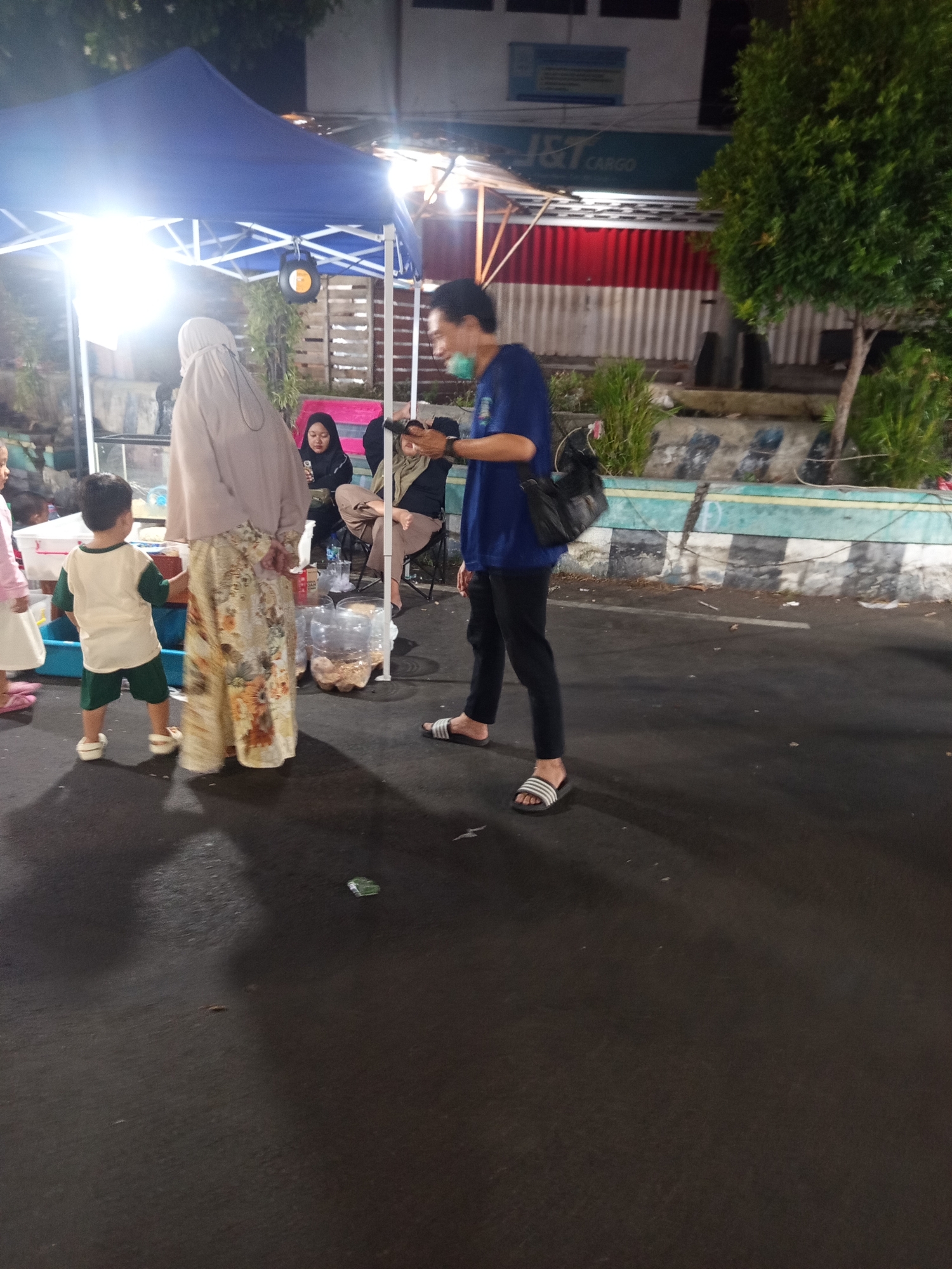 The image captures an evening scene on a street in Ardirejo, Indonesia, likely at a market or event. In the foreground, a man wearing a blue t-shirt, black pants, and striped sandals is walking. He has a black bag slung over his shoulder and is wearing a light-colored face mask. To his left, a child in a white and green outfit stands facing away from the camera. Behind the child is a woman wearing a beige hijab and a long, patterned dress, and another woman in a black hijab is seated. A blue tent is set up behind them, illuminated by bright lights. In the background, a building with a red and white striped facade and a sign that reads "J&T CARGO" is visible, along with trees and a street curb. The ground is paved asphalt.