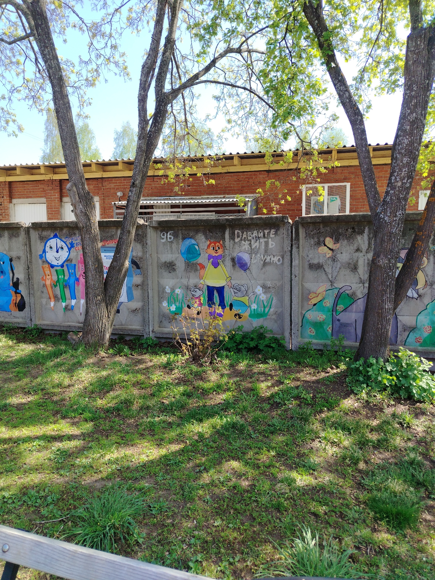 The scene depicts a painted concrete fence in what appears to be a park or public space. The fence is decorated with colorful children's drawings, including cartoon characters, animals, and flowers. The number "96" is written on one section of the fence, and a message in Russian is visible on another section. Behind the fence is a red brick building with a metal roof. Trees with sparse green foliage are in the foreground, partially obscuring the fence. The ground is covered in green grass with patches of dirt and shadow. The lower part of a wooden bench is visible in the foreground. This image was taken in the city of Noviy, Russia.