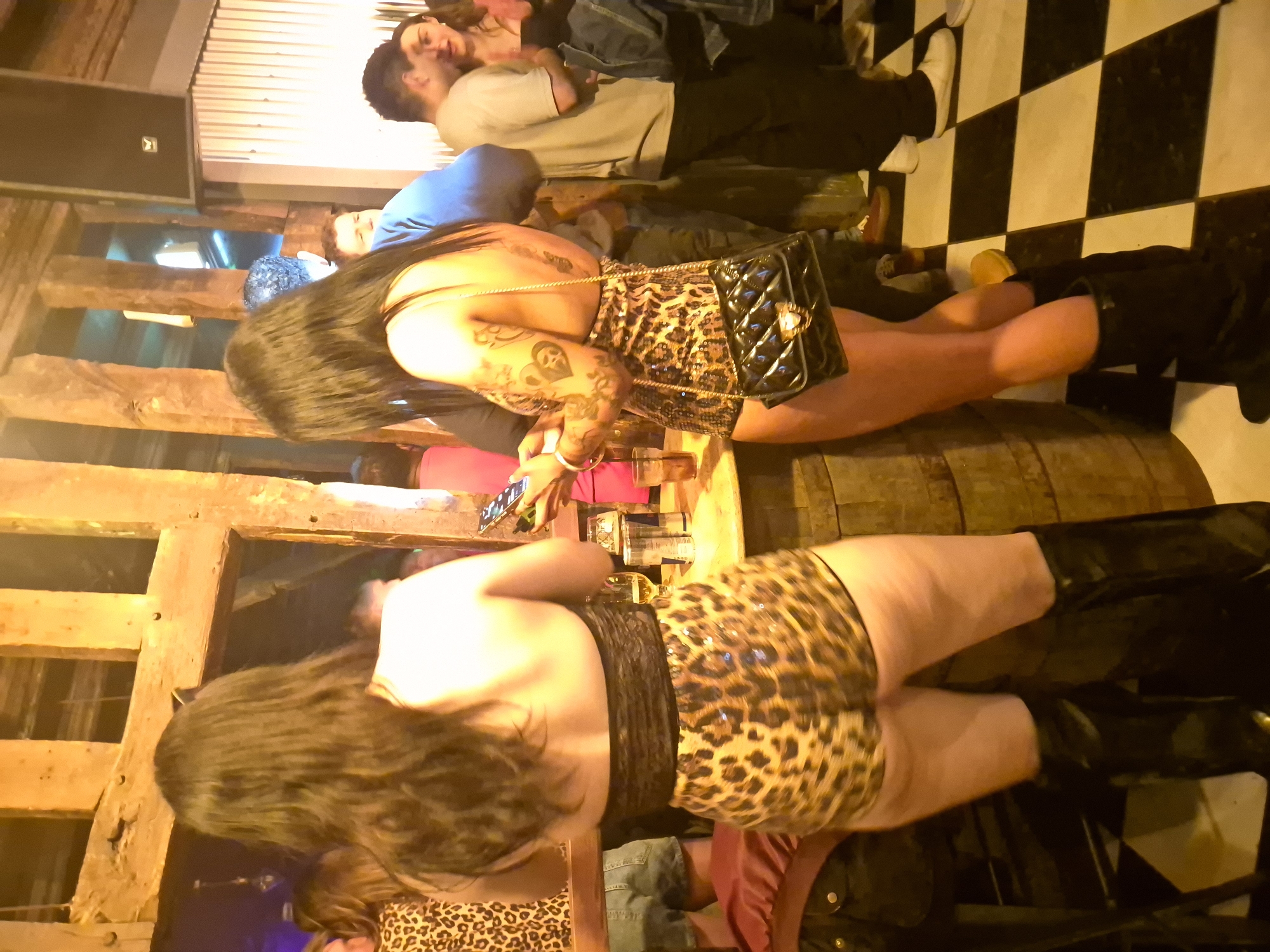 The image depicts an indoor scene, possibly a bar or club, based on the exposed wooden beams, mixed flooring including checkered tiles, and a partially visible metal corrugated ceiling. Multiple people are present. One woman is prominently displayed in a leopard print outfit and high boots, seemingly posing for a photograph taken with a mobile phone. Another woman, similarly dressed, is also in view. Several other individuals are in the background, including a couple embracing. The location is inferred to be in West Suffolk, United Kingdom.