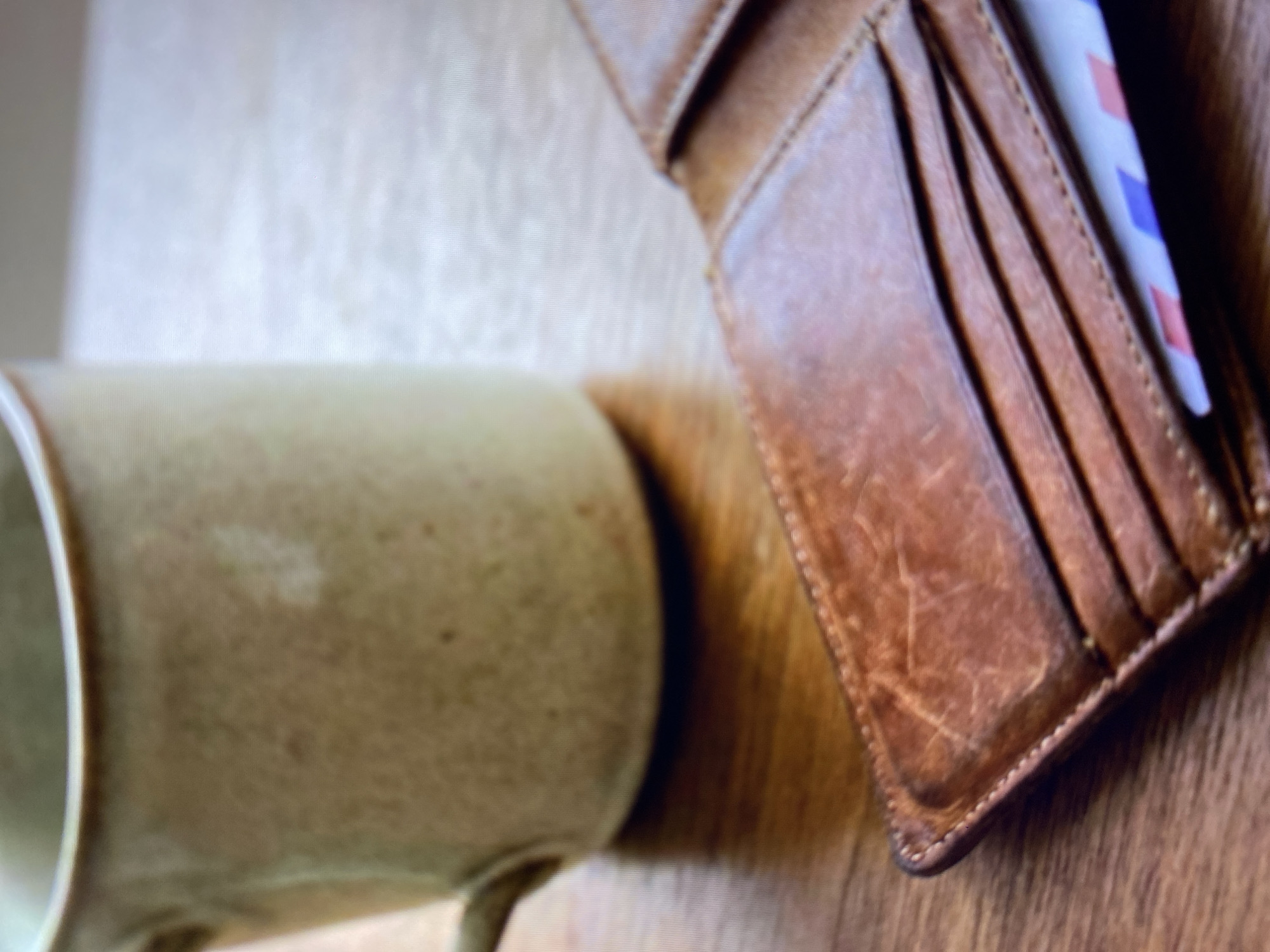 A close-up, eye-level shot shows a rustic, light brown ceramic mug with a handle on the left, positioned on a wooden surface. To the right of the mug is an open, distressed brown leather wallet. Several card slots are visible within the wallet, with a portion of a card or paper showing a pattern of pink and blue stripes peeking out from the bottom. The background is a blurry, neutral tone. The scene suggests a casual, domestic setting, possibly a desk or table, and does not depict any specific location in Kuta Selatan, Indonesia.