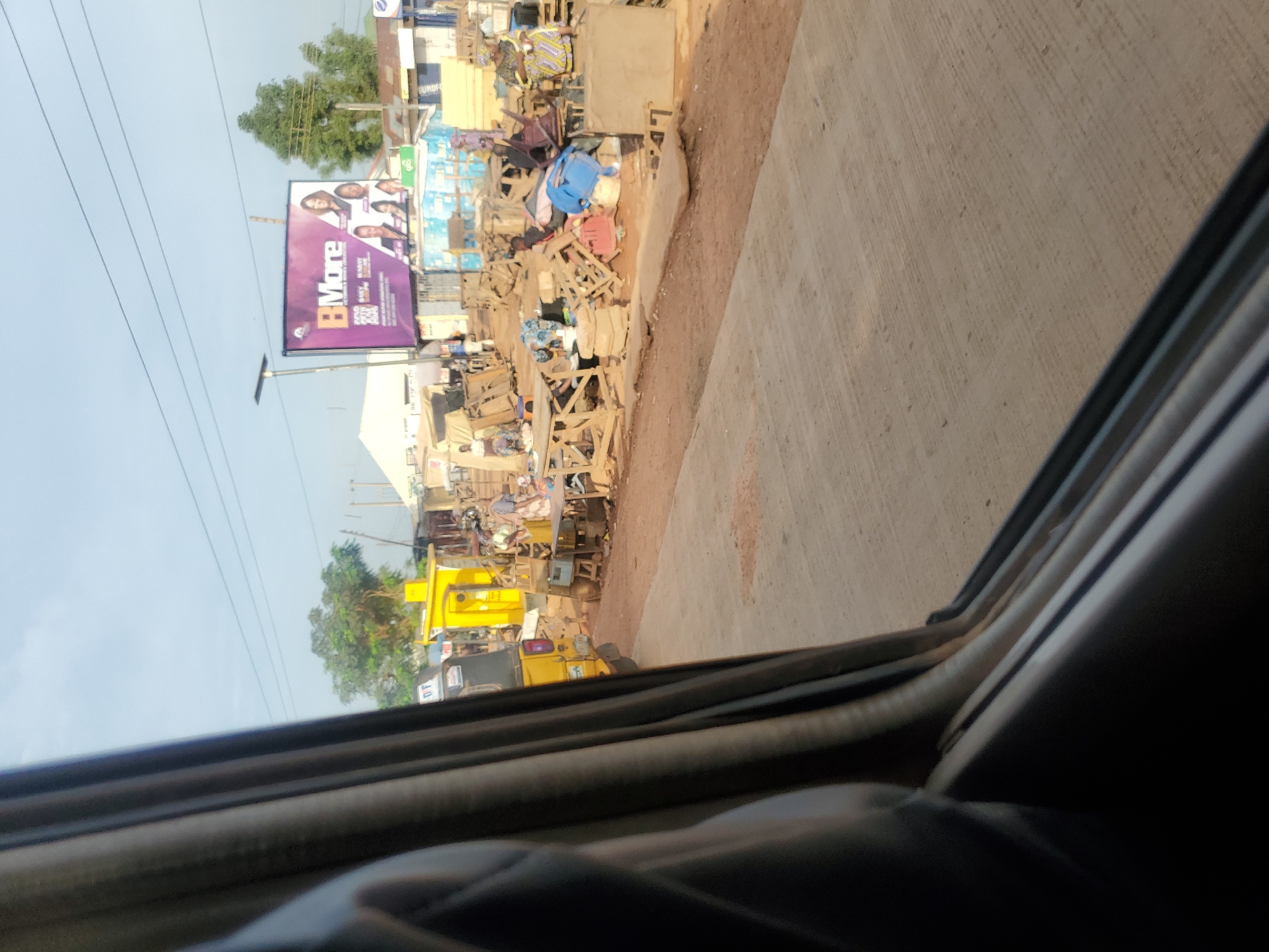 The image depicts a street scene in Goni Gara, Nigeria. In the foreground, the dark interior of a vehicle is visible, with the edge of a window frame and a black fabric lining the left side of the frame. The road surface in front of the vehicle is paved concrete.
Beyond the road, the scene opens up to a bustling market or roadside trading area. A yellow kiosk or small building is prominent on the left, with a portion of a yellow motorized tricycle parked nearby. Various stalls and makeshift structures made of wood and corrugated metal are set up along the roadside, displaying goods and seating areas. Several wooden chairs and tables are stacked and arranged.
People are visible in the market area, some appearing to be vendors or shoppers. One woman in a colorful patterned dress stands near a stall on the right. Other individuals are engaged in activities related to the stalls, such as arranging items or sitting. A large billboard is situated in the background, advertising "BMore" with images of women. The environment appears to be outdoors, with trees and some buildings in the distance, under a hazy sky. Power lines are visible stretching across the upper portion of the image. The overall impression is one of a vibrant, informal commercial district.