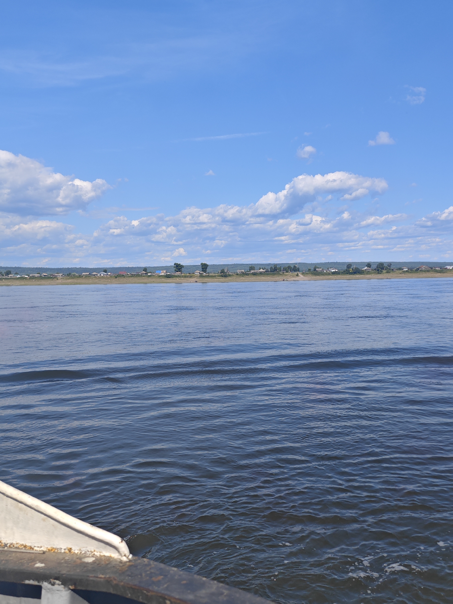 The image depicts a wide, calm river under a bright blue sky with scattered white clouds. The river's surface shows gentle ripples. In the distance, across the river, a low-lying landscape is visible, characterized by a line of trees and a small settlement of houses. The settlement appears to be a village with scattered buildings, some with red roofs. The overall impression is one of a serene rural or semi-rural environment. This scene is consistent with the geography of Lesosibirsk, Russia, a city located on the banks of the Angara River, a tributary of the Yenisei.