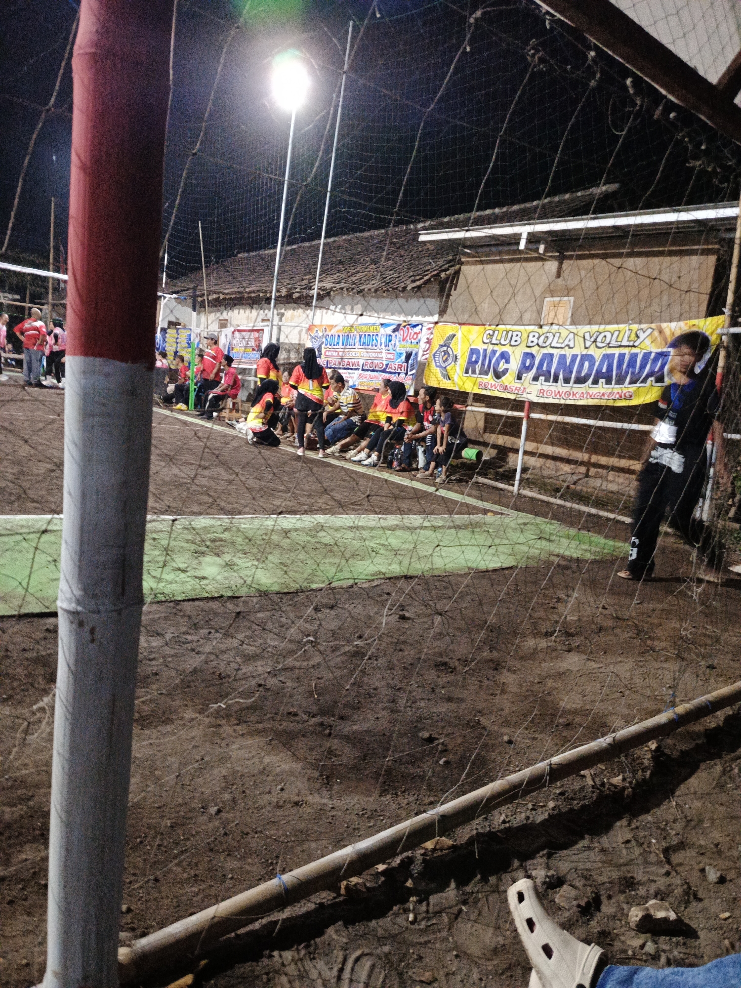 The image depicts an outdoor volleyball court at night, likely in Rowokangkung, Indonesia, illuminated by a bright overhead light. A volleyball net is prominently featured in the foreground, with a red and white striped pole to its left. In the background, a banner is visible, reading "CLUB BOLA VOLLY RUC PANDAWA" and also "OPEN TURNAMEN BOLA VOLI KADES CUP". Spectators, some wearing red and yellow, are seated on benches or the ground along the edge of the court. A few people are standing or moving around the court area. The ground appears to be dirt. A person in dark clothing is leaning on the net support on the right side of the frame.