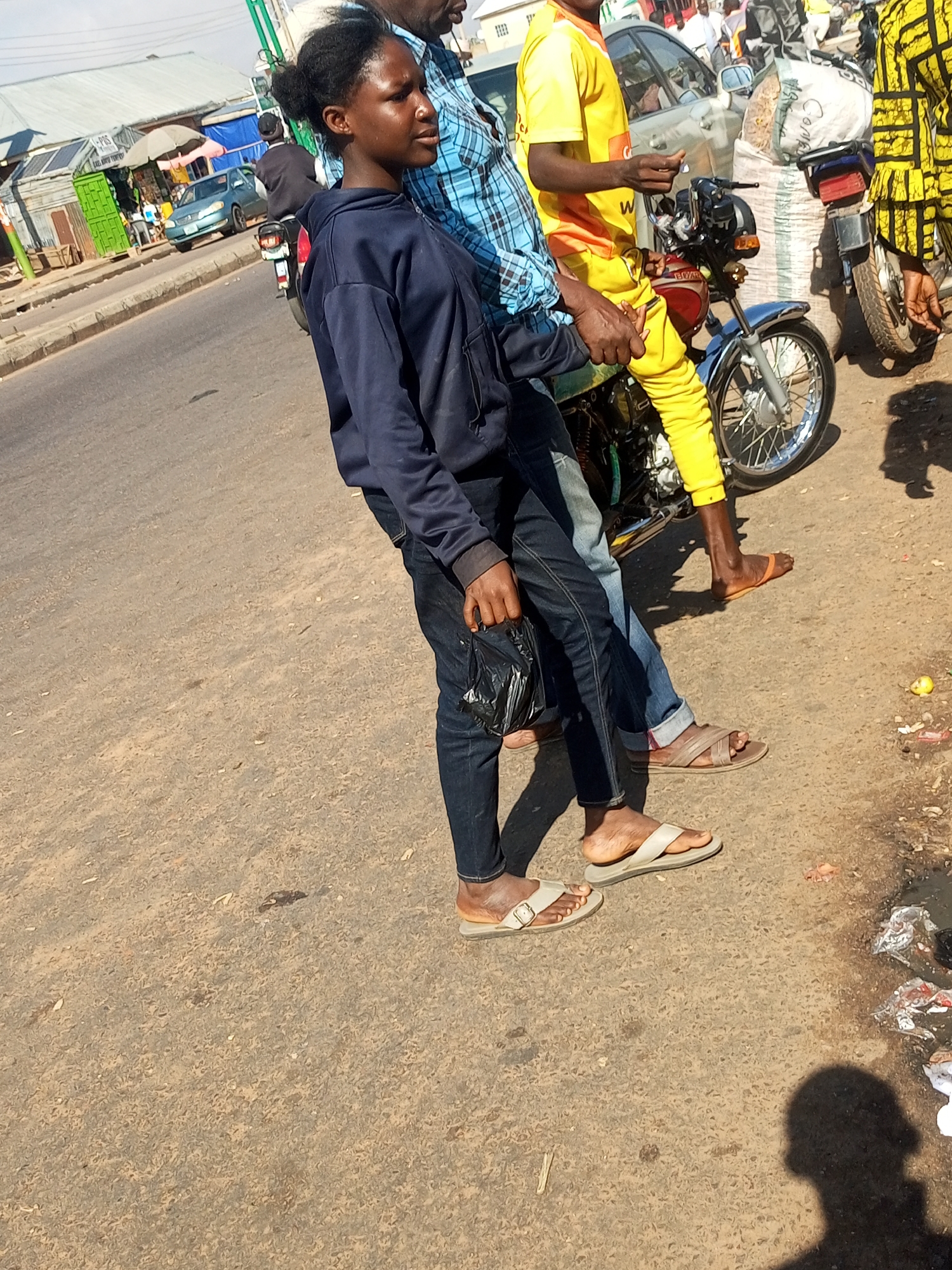 In Tempure, Nigeria, a young woman, dressed in a dark blue hoodie and jeans, stands on a dusty road next to a motorcycle. She holds a black plastic bag in her right hand. Behind her, a man in a blue checkered shirt and another man in yellow attire are partially visible. The man in yellow is seated on the motorcycle. A large sack of what appears to be grain or feed is propped up behind the motorcycle. In the background, a light blue car is parked on the side of the road, and a market stall with various goods is visible. The scene suggests a bustling street environment with commercial activity.