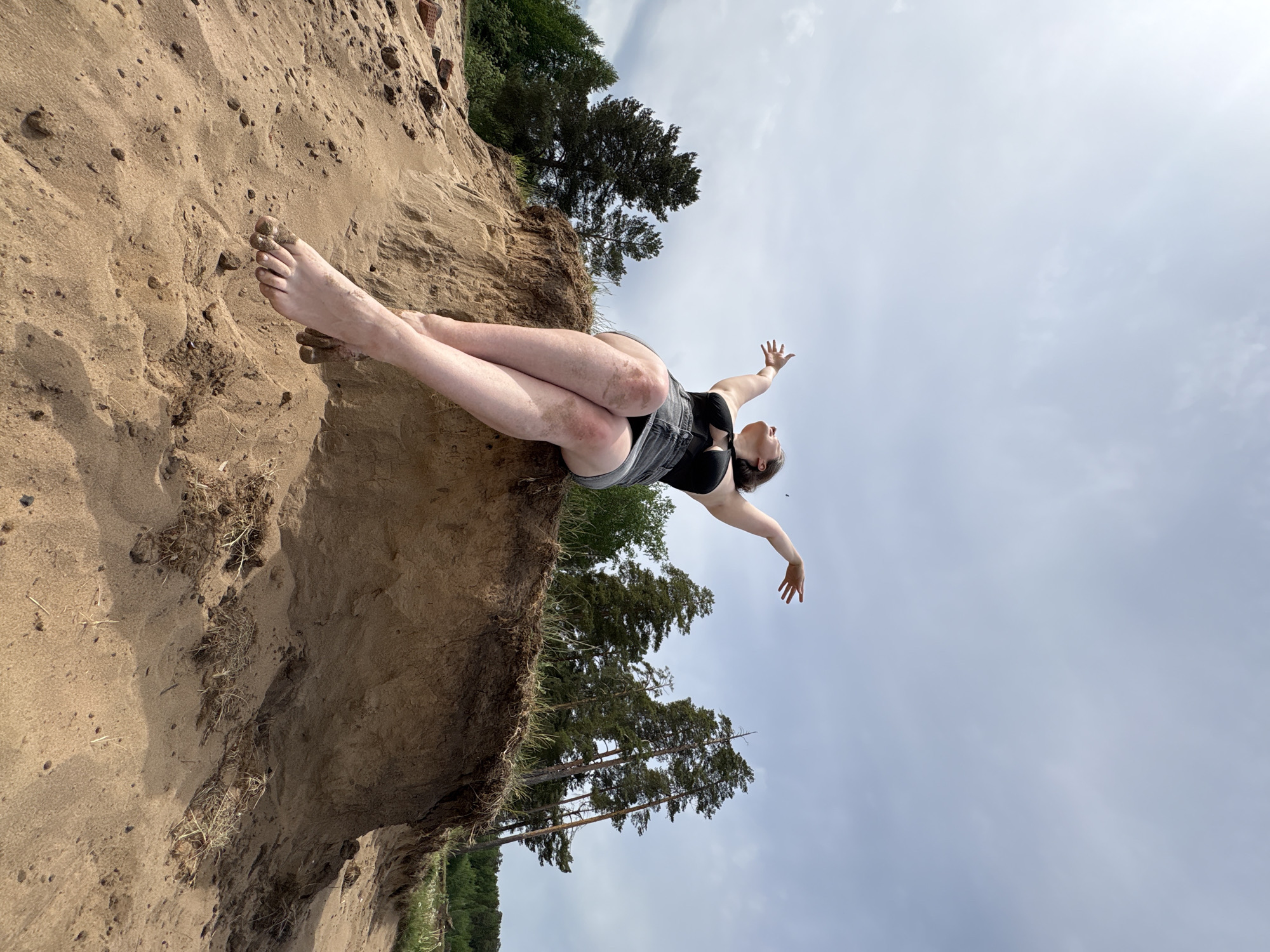 The image shows a woman lying on a sandy cliff in Chaikovsky, Russia, with her legs resting on the sand and her arms outstretched towards the sky. She is wearing a black top and gray shorts. The background includes trees and a cloudy sky.