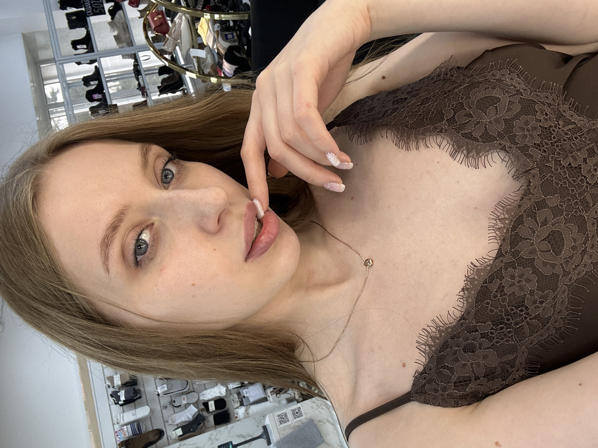 A young Caucasian woman with long, light brown hair, blue eyes, and light skin is the main subject of this image. She is wearing a brown lace-trimmed camisole and a delicate gold necklace. Her right hand is near her face, with fingers lightly touching her lips. Her fingernails are painted with a light, neutral color with subtle patterns.
The background shows a store environment, with shelves stocked with various items. The location appears to be in the city of Chaikovsky, Russia, based on the presence of Cyrillic script in the background.