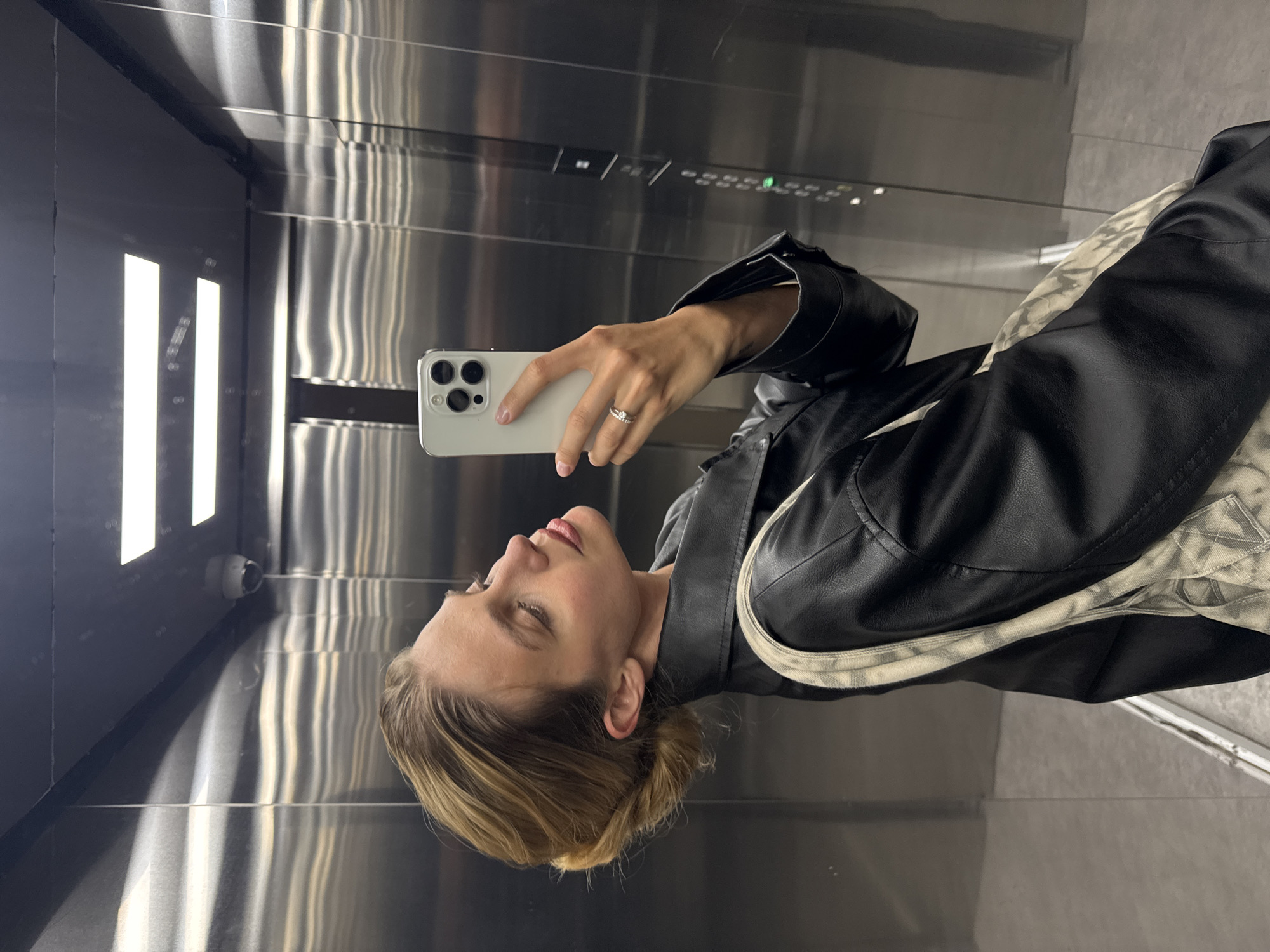 A person stands in an elevator, taking a selfie with a white smartphone. The individual is wearing a black leather jacket with a light-colored lining. The elevator's interior is predominantly metallic, with visible buttons and a security camera.