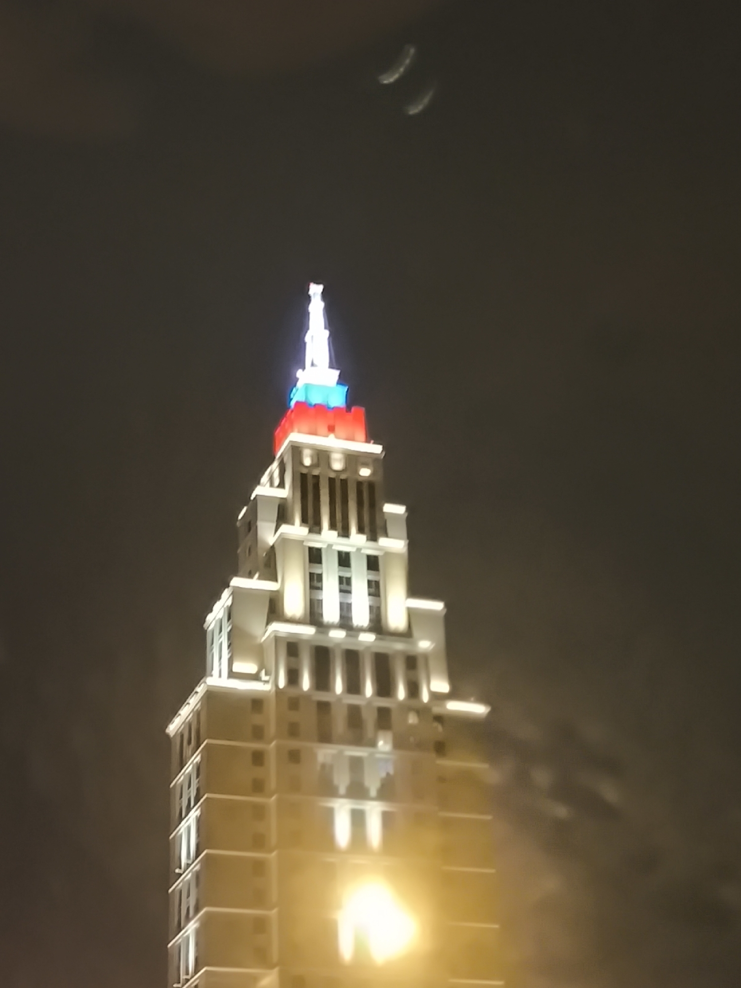 At night, a tall building in Moscow, Russia, is illuminated. The top of the building is lit with white, red, and blue lights. The building is lit with yellow lights. The sky is dark and cloudy.
