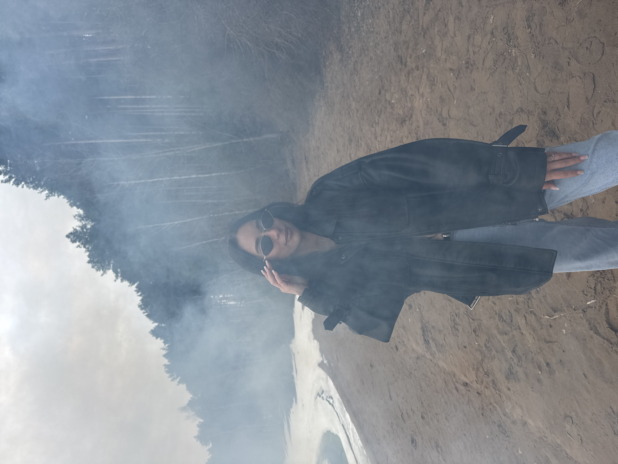 The image depicts a woman standing outside near a body of water in Chaikovsky, Russia. She has long dark hair and is wearing sunglasses, a black leather jacket, and light-wash jeans. She is facing the camera and has one hand raised near her face. There is a noticeable fog or smoke in the air, obscuring a view of trees in the background. The ground appears to be sandy or dirt. The water is visible in the lower portion of the image, and the background is somewhat blurry, possibly due to the smoke or fog.