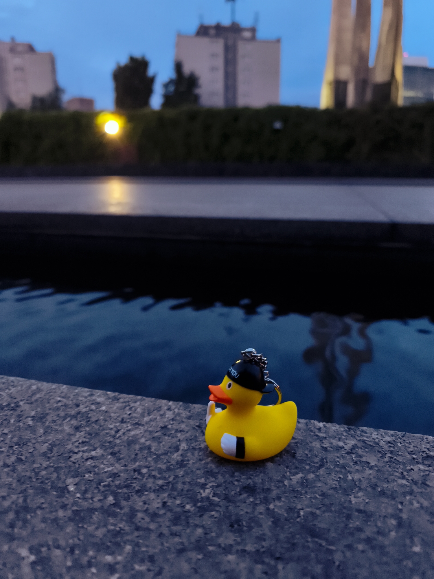 A yellow rubber duck toy with a black hat and a keychain is positioned on a stone ledge beside a body of water. The duck is facing left, with its beak slightly open. The hat has "OK YOU" written on it in white letters. In the background, out of focus, are buildings, trees, and a lit lamp. The water appears to be a canal or river, reflecting the dim twilight sky and surrounding structures. The scene is set in Gdańsk, Poland, near the Motława River.