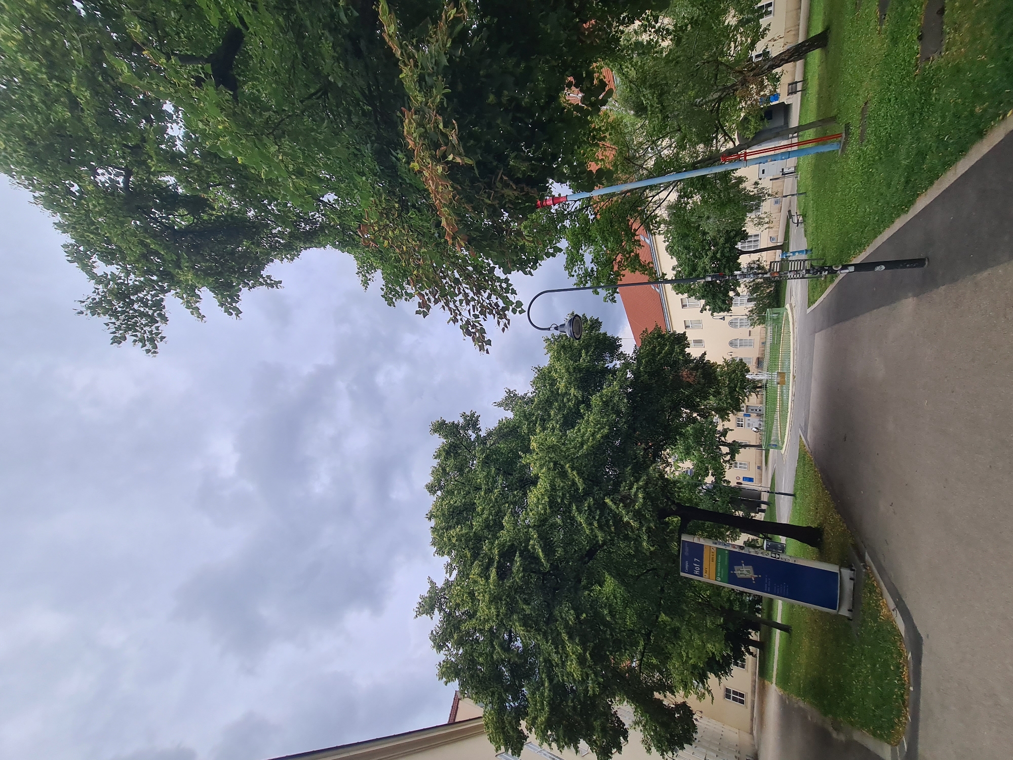 The image depicts an outdoor scene with a pathway leading into a campus area. In the foreground, there is a paved walkway bordered by green grass. To the left, a tall blue sign with text and a diagram is partially visible, indicating "Campus Hof 7" and a map of the area. A large, leafy green tree dominates the left side of the frame, with part of a cream-colored building visible behind it.
In the mid-ground, the pathway curves into a central courtyard. More trees with dense foliage are scattered across the lawn. A lamppost with a curved arm is visible. To the right, another building with a red-tiled roof and white windows can be seen. Two colorful poles, one blue and one red, stand amidst the trees. Benches are placed on the grass. In the background, a circular fountain with green trim is active, with water spraying upwards. The sky is overcast with gray clouds. The overall setting appears to be a university campus.
Based on the "Campus Hof 7" sign and architectural style, this location is likely part of the University of Vienna's Altes AKH (Old General Hospital) campus.