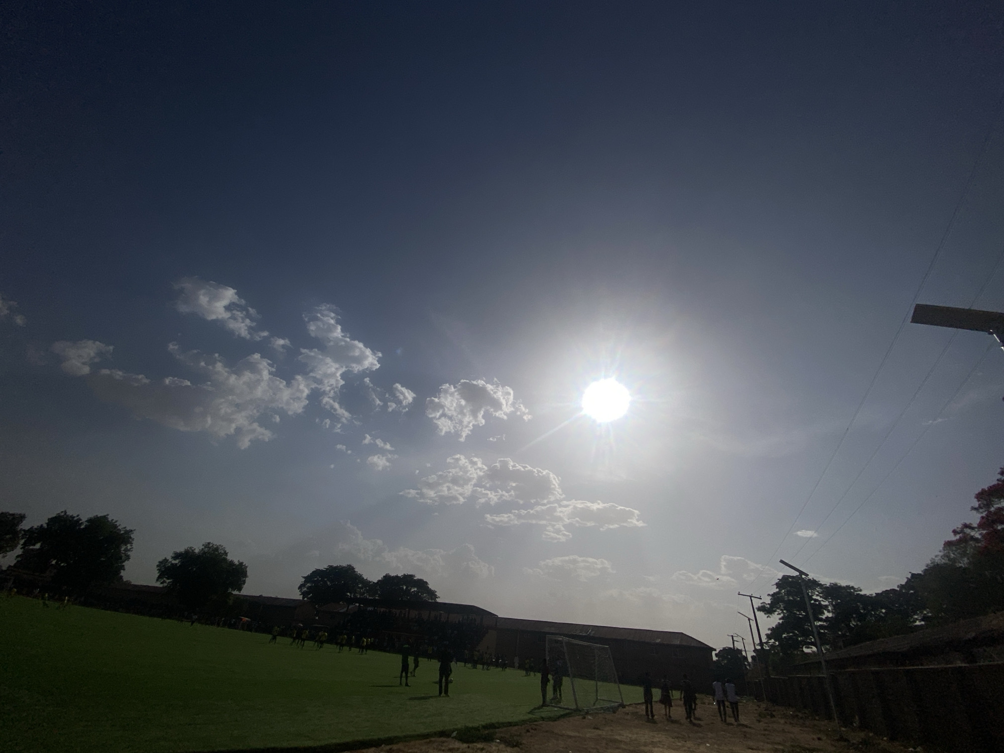 The image depicts a sunny day in Tudun Wada, Nigeria, with a bright sun obscured by light clouds. A green field occupies the foreground, where several people are engaged in activities resembling a soccer practice. A goalpost is visible on the field. In the background, there is a building and trees. Power lines and poles are visible on the right side of the frame. A fence runs along the edge of the field. The overall scene suggests an outdoor activity occurring on a field in Tudun Wada.