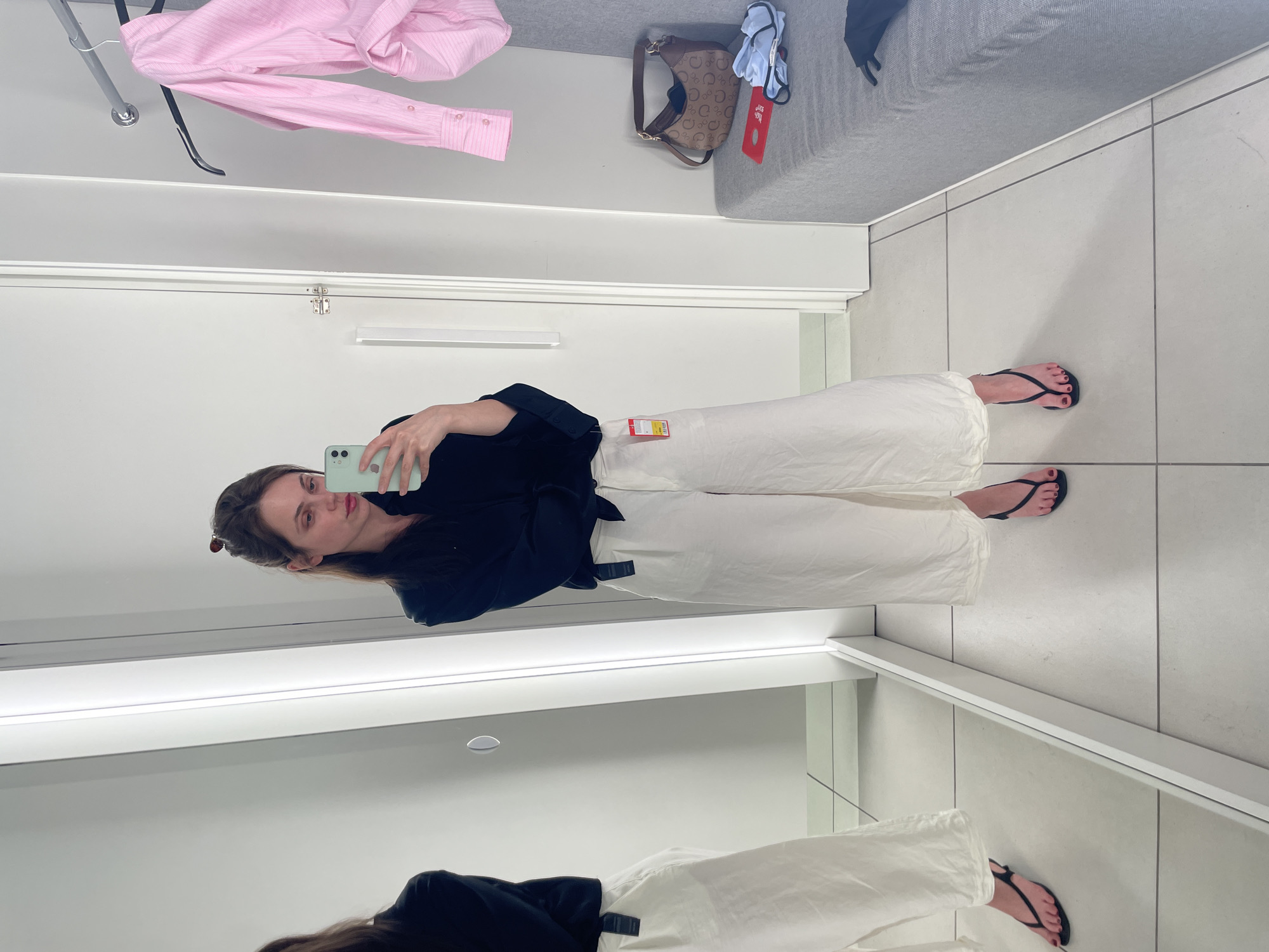 A woman is taking a mirror selfie in a brightly lit fitting room. She is wearing a black long-sleeved blouse and wide-leg white pants, accessorized with black thong sandals. Her hair is pulled back in a bun, and she is holding a light green smartphone. To her right, a pink striped shirt hangs on a clothes rack, and a patterned handbag rests on a gray bench next to some folded clothing. The fitting room has white tiled floors and walls, with a full-length mirror and a doorway visible. The scene appears to be in a clothing store, likely in Vecsés, Hungary, given the provided context.