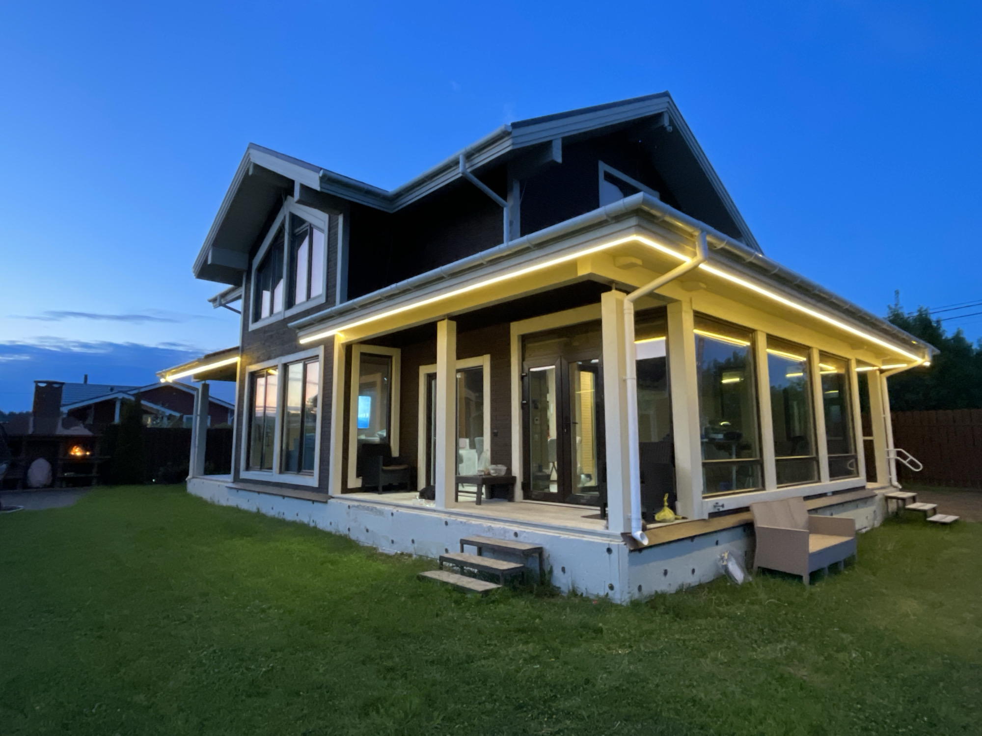 The image depicts a modern, two-story house in Solov'evka, Russia, taken during twilight. The house features a dark brown exterior with white trim and large windows. A porch with white columns runs along the front and side, illuminated by a string of lights. The house sits on a concrete foundation with exposed insulation. In front of the house is a green lawn, and a wooden fence is visible in the background on the right. To the left, there is an outdoor fireplace with a chimney. A chair is on the porch.