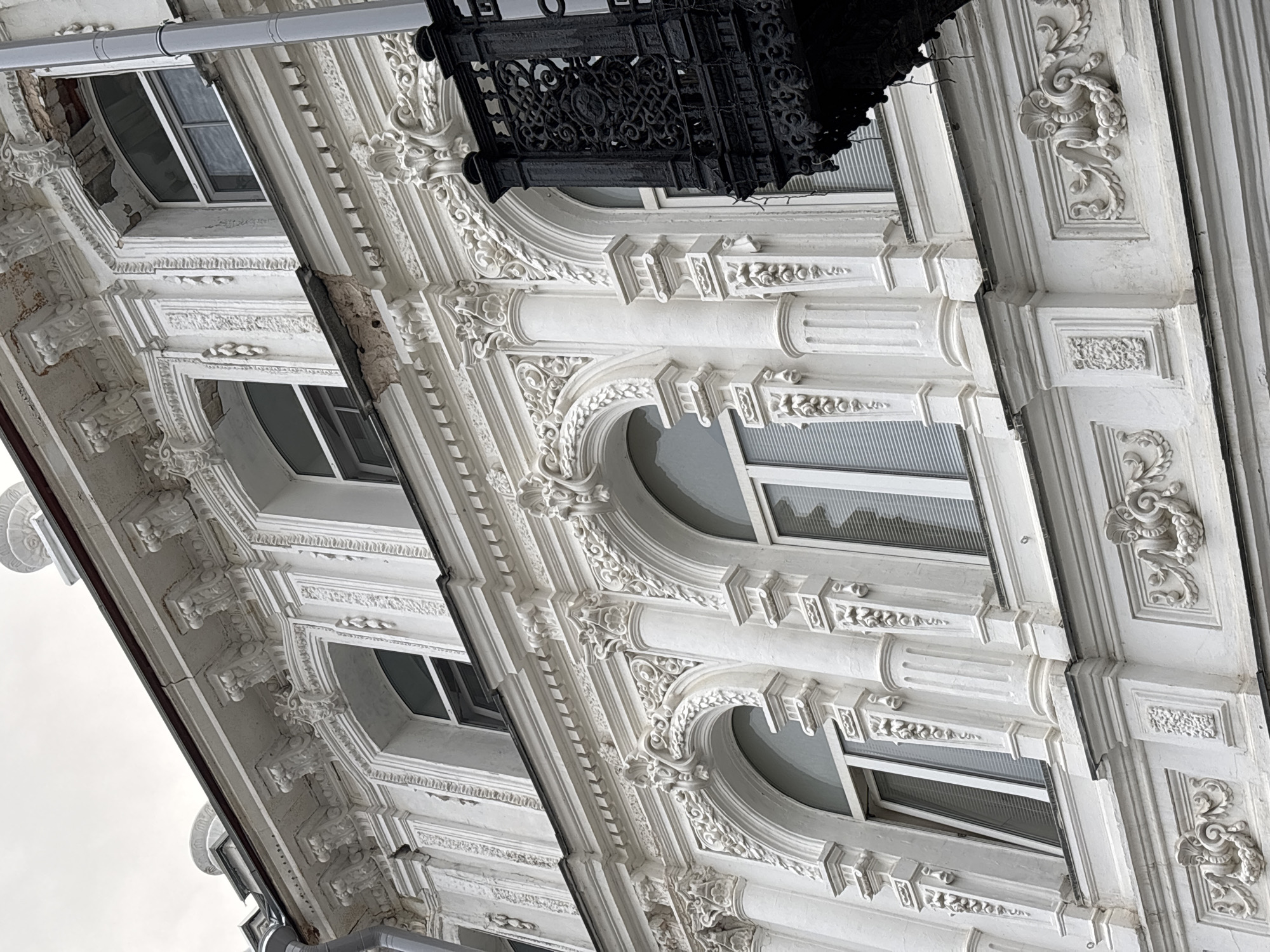 The image shows a close-up, low-angle view of the ornate facade of a building in Nizhny Novgorod, Russia. The building appears to be old, with weathered white stucco and detailed architectural elements. The facade features multiple arched windows with white frames and blinds. Intricate decorative moldings, including floral motifs and classical columns, adorn the exterior. A black wrought-iron balcony with a decorative railing is visible on the right side of the image, extending from an upper floor. The sky above is overcast. The building's condition suggests a need for restoration, with some areas of chipped stucco and exposed brickwork visible.