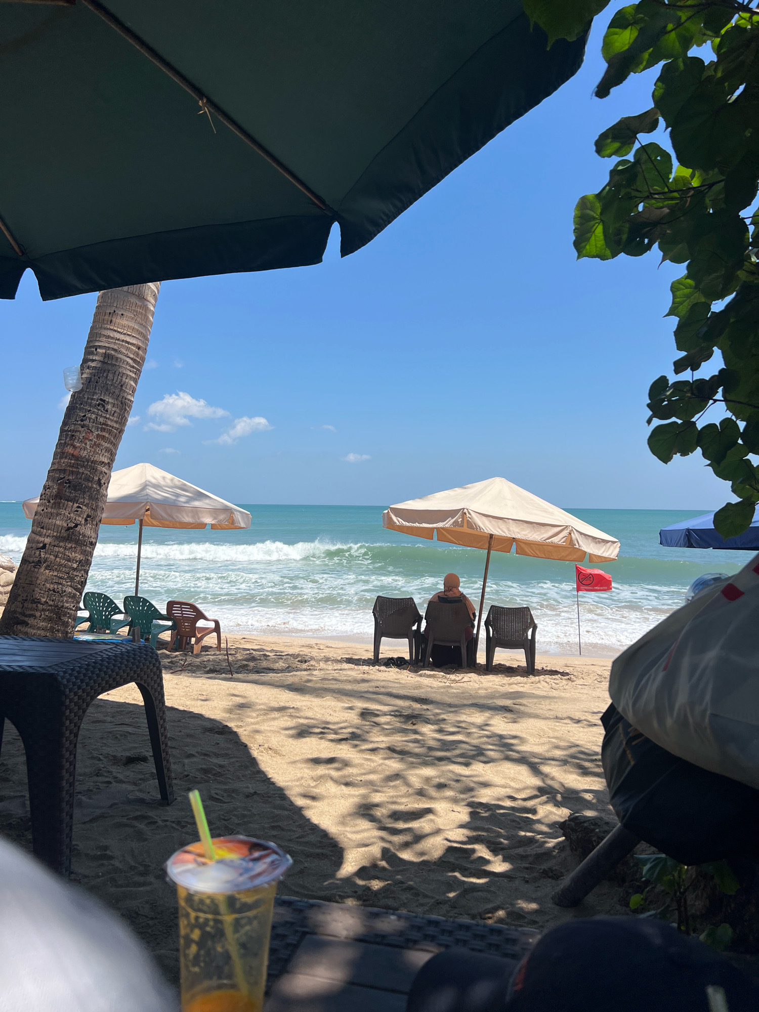 The image depicts a serene beach scene in Kuta, Indonesia, bathed in bright daylight. In the foreground, a large, dark green umbrella casts a shadow over the sandy shore, obscuring part of a table. To the left, a palm tree trunk stands tall, with a plastic bottle affixed to it. Further back, two beige beach umbrellas provide shade for chairs arranged on the sand.
In the mid-ground, a person sits facing the ocean, with two empty chairs beside them. A small red flag with white text is planted in the sand near the water's edge, indicating a warning or restriction. The turquoise ocean stretches to the horizon, with gentle waves breaking on the shore. To the right, foliage from a tree partially frames the scene, and another umbrella, this one blue, is visible in the distance.
In the immediate foreground, a clear plastic cup filled with an orange beverage and a green straw is placed on a surface that appears to be a table. The sand is a light tan color, marked with shadows from the umbrellas and trees. The sky is a clear, vibrant blue with a few scattered white clouds. The overall impression is one of a tranquil beach day.