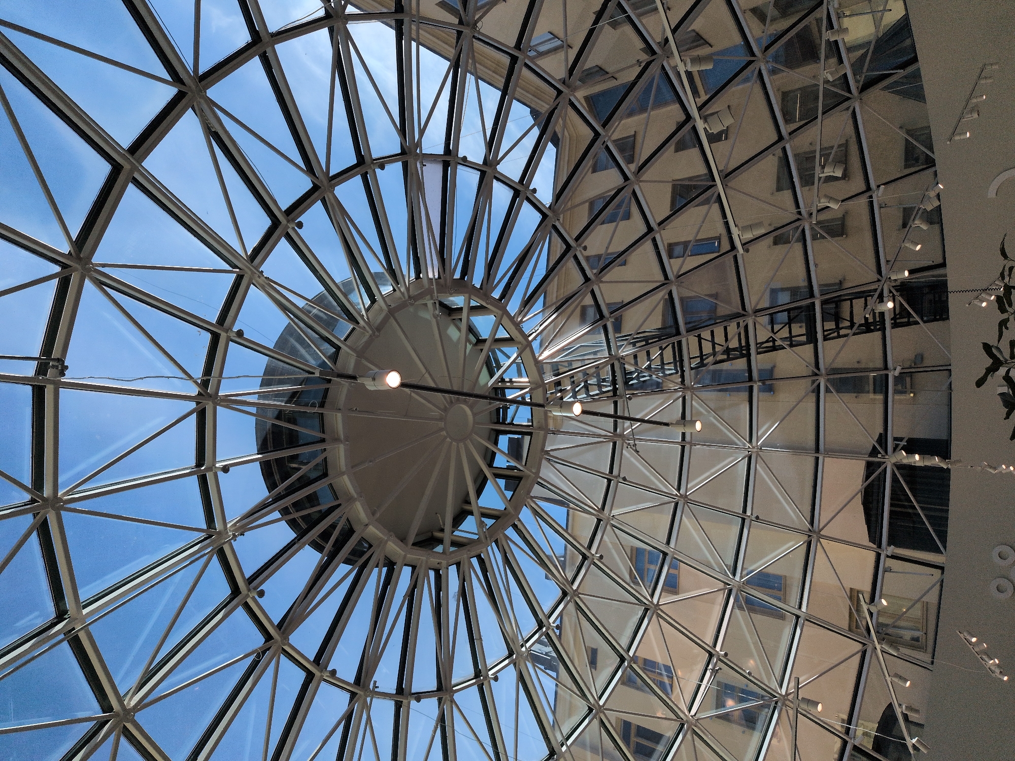 The image shows the interior of a building with a geodesic dome-shaped glass ceiling. The ceiling is supported by a network of white metal beams that form triangular patterns. Through the glass, a clear blue sky with some wispy clouds is visible. Reflections of a building with multiple windows are visible on the glass panels. A black metal fire escape or maintenance ladder is visible ascending within the dome. Several spotlights are attached to the ceiling structure and to wires hanging down. In the lower part of the image, a light-colored curved wall is visible, with some plants and hanging light fixtures. This scene is located at the Forum shopping center in Helsinki, Finland. There are no people visible in the image.