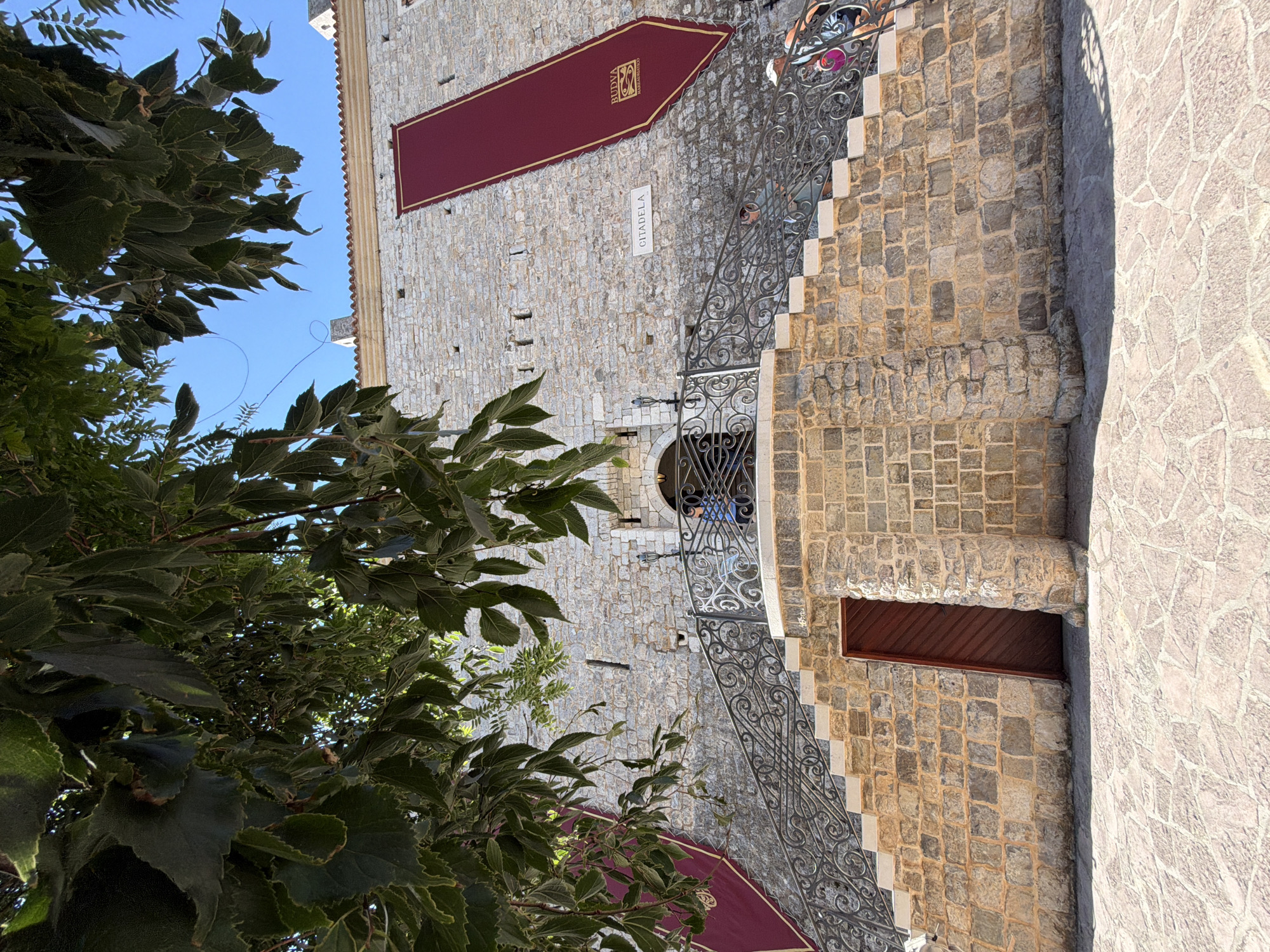 The image shows a stone fortress in Budva, Montenegro, identified by a sign as "CITADELA". A wide, stone staircase with ornate metal railings leads up to an arched entranceway in the fortress wall. Two large, burgundy banners with gold trim and insignia hang vertically on the fortress wall. Green foliage from a tree hangs over the top of the scene, partially obscuring the clear blue sky. The ground in front of the staircase is paved with irregular stones. A wooden door is visible at the base of the staircase, set into a stone wall. People are visible on the staircase and near the entrance.