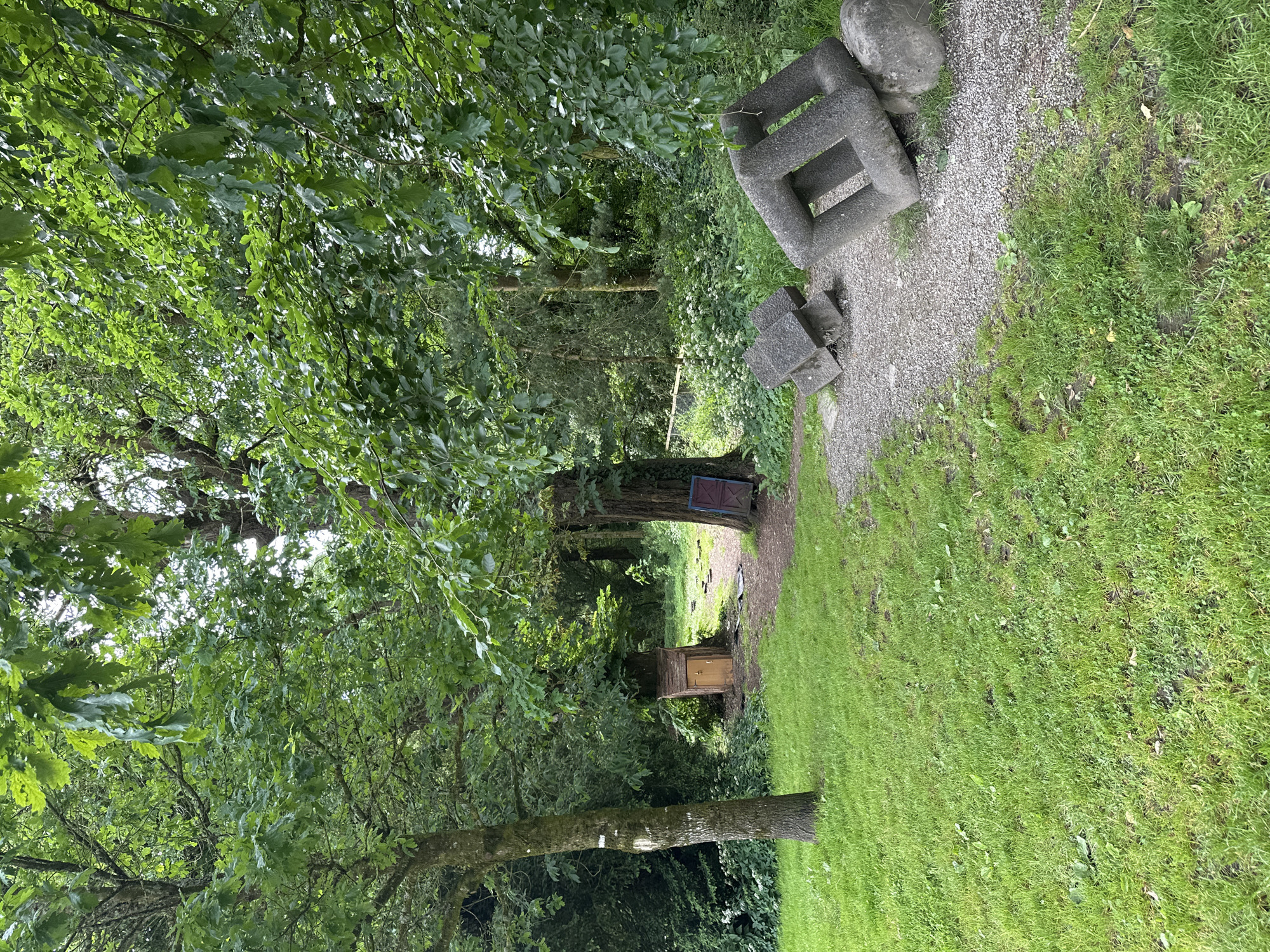 An outdoor scene features a winding gravel path through a wooded area with lush green grass. On the left, a tree trunk with a white marking stands prominently. Further down the path, a small, wooden structure resembling a shed or miniature house with a single door is visible amongst the trees. To the right of the path, near the foreground, are two abstract geometric sculptures made of grey stone. The sculptures appear to be interconnected block-like shapes, with one resting on a rounded stone. The surrounding environment is dense with various trees and foliage, suggesting a natural or park-like setting. The lighting suggests it is daytime.