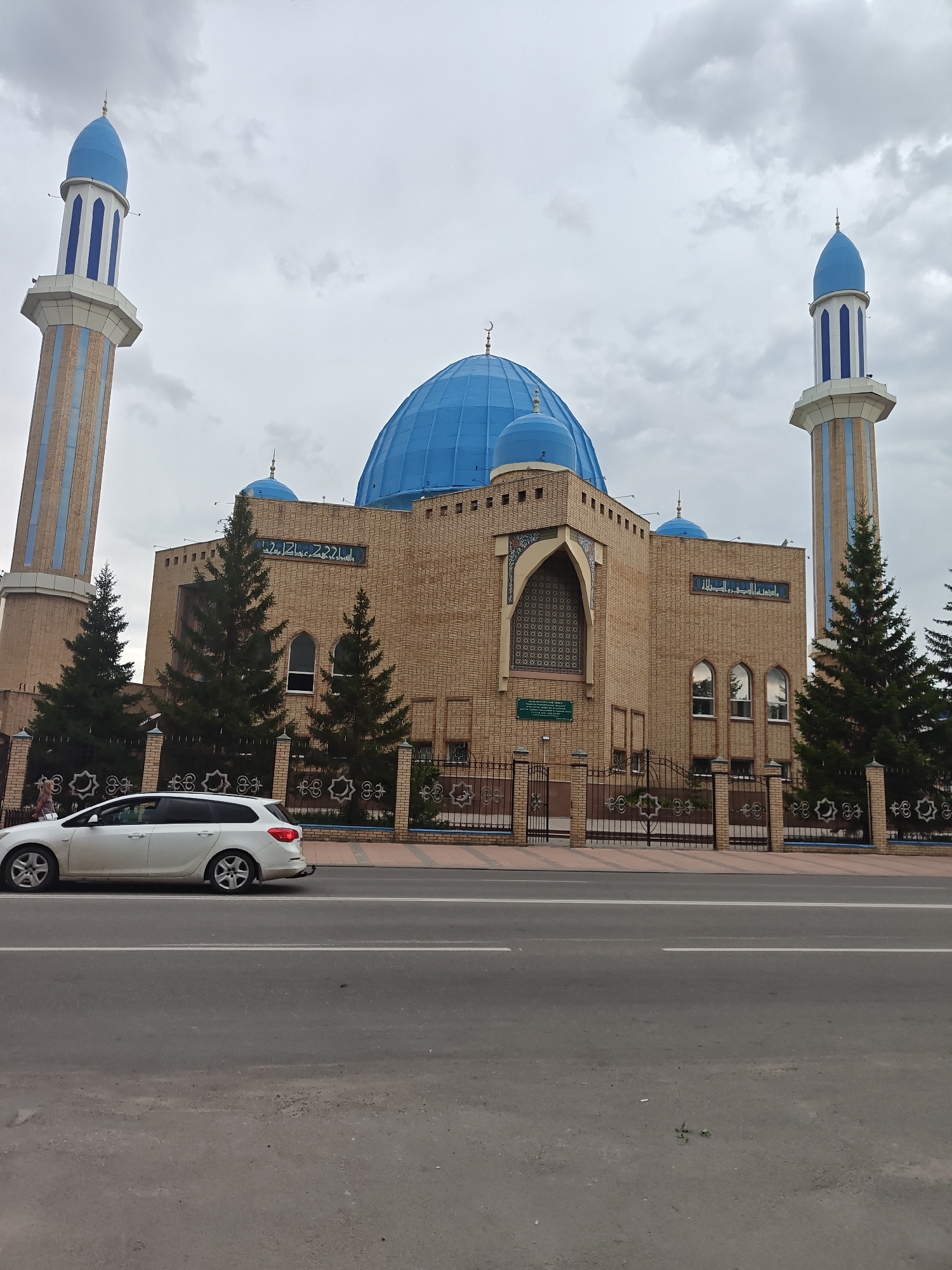 The image depicts the North Kazakhstan Regional Mosque in Petropavl, Kazakhstan. The mosque features two tall, slender minarets with blue and white striped detailing and blue conical tops, flanking a central building with a large blue dome. The building is constructed from light brown brick. In front of the mosque, there is a black metal fence with decorative elements and a walkway. Several evergreen trees are planted along the fence. A white station wagon is driving on the road in the foreground, and a pedestrian is walking on the sidewalk near the fence. The sky is overcast with grey clouds.