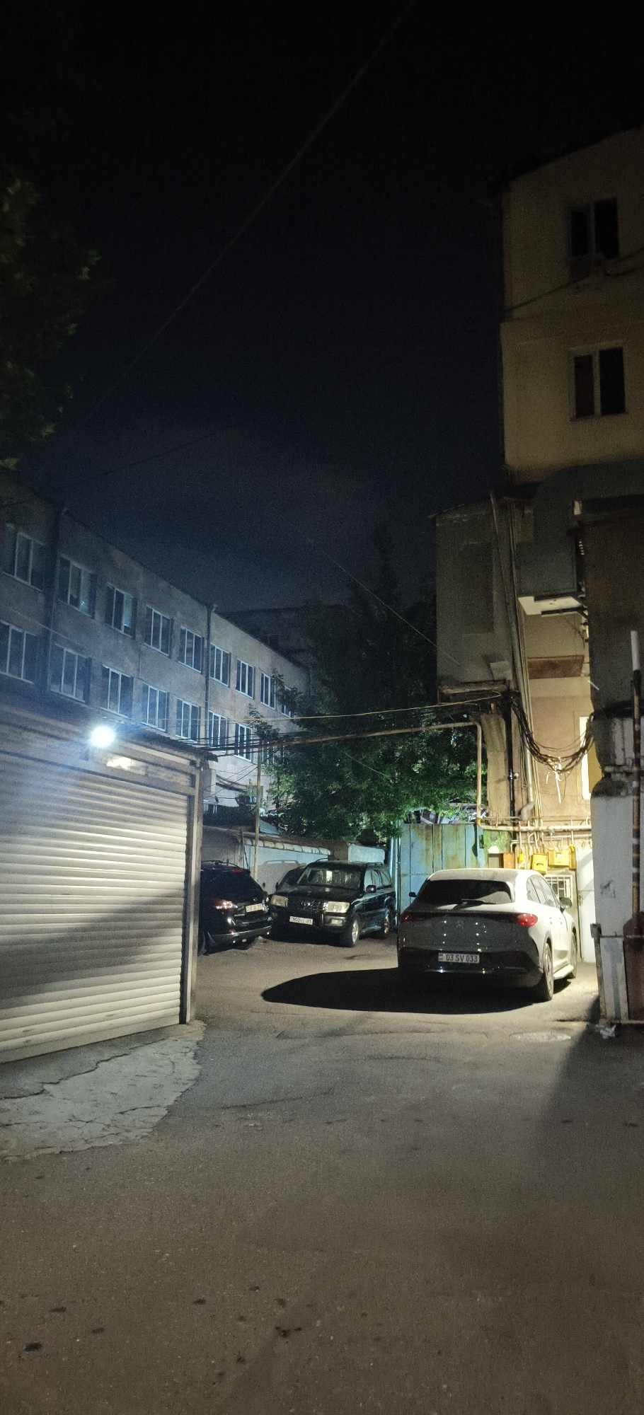 This image depicts a nighttime scene in an urban environment, likely a courtyard or alleyway between buildings. In the foreground, asphalt pavement is visible, with some debris and cracks. To the left, a large, ribbed garage door, likely made of metal, is illuminated by a bright overhead light. Several cars are parked in the area.
On the left, a dark-colored SUV is parked, partially visible. Next to it, a black SUV with a license plate is also parked. To the right of these, a white SUV with a license plate is positioned, facing away from the viewer.
In the background, a multi-story building with numerous windows lines the left side of the scene. The building appears to be residential or office space. To the right, another building is visible, with architectural features and external wiring. Trees with dense foliage are present, casting shadows in the artificial light. The overall impression is one of a utilitarian, possibly residential, urban space at night.