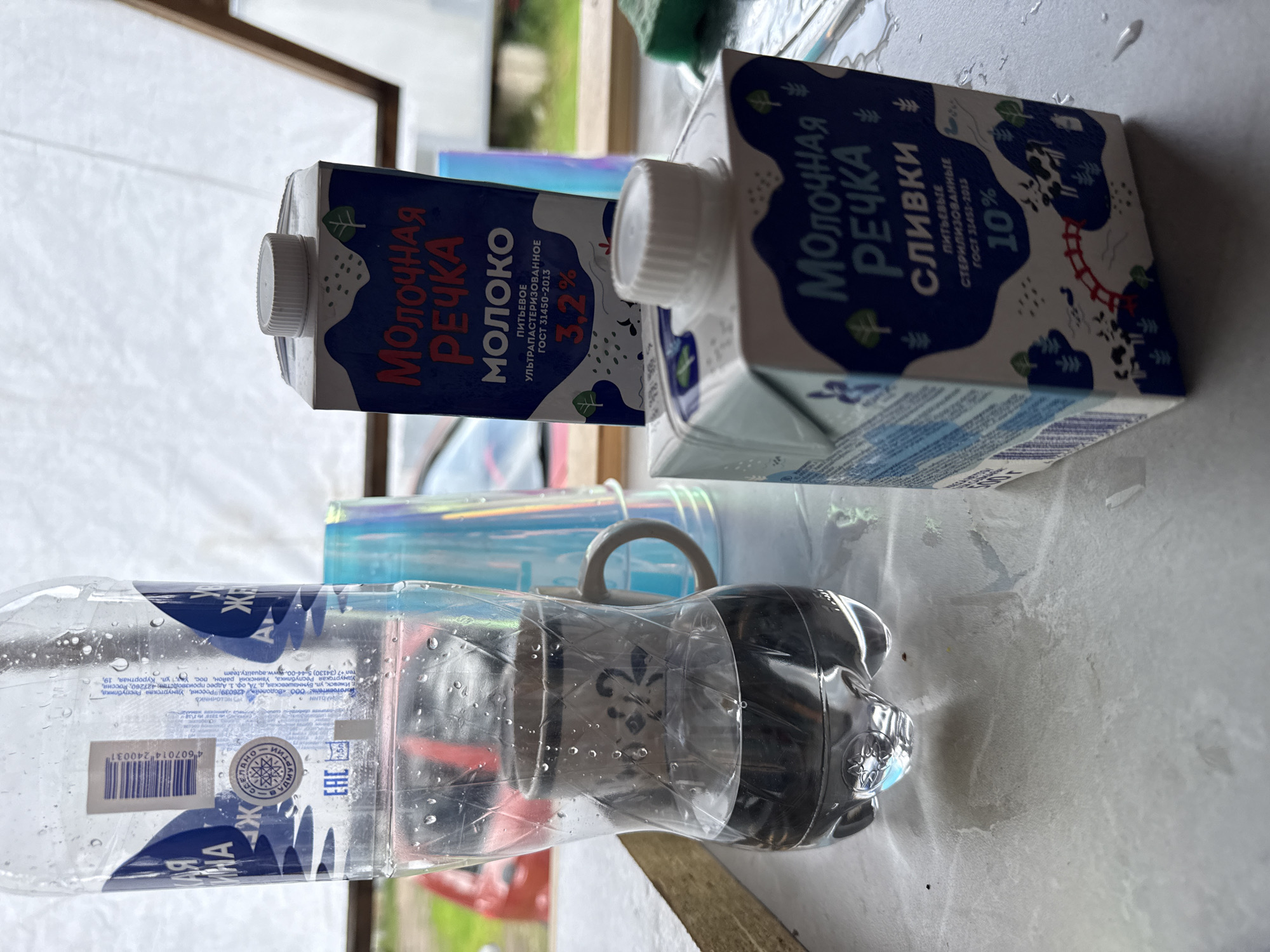 The image shows a close-up of several beverage containers and a mug on a light-colored surface. In the foreground, there are two small cartons of dairy products. The one on the right is labeled "МОЛОЧНАЯ РЕЧКА СЛИВКИ 10%" (Milk River Cream 10%). To the left of this, partially obscured, is another carton of "МОЛОЧНАЯ РЕЧКА МОЛОКО" (Milk River Milk) with "3.2%" visible. Behind these cartons, a clear plastic bottle is filled with a dark liquid. Inside the bottle, a ceramic mug is placed. The background shows what appears to be a slightly out-of-focus outdoor scene with some foliage and a red object, possibly a vehicle. The setting is likely outdoors or in a space with natural light, given the lighting and the presence of outdoor elements. The presence of water droplets on the plastic bottle suggests humidity or condensation. The scene is reminiscent of a casual outdoor setting, possibly a picnic or a backyard gathering. There are no people visible in the image. The location is identified as Chaikovsky, Russia, as indicated by the label on the water bottle, which includes an address in Chaikovsky.