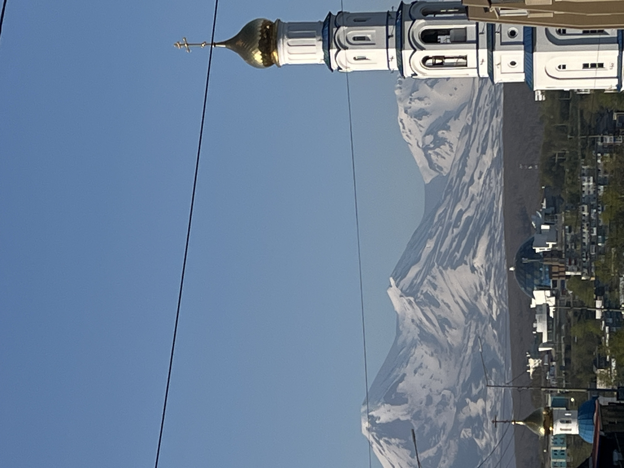 The image depicts a cityscape with a prominent white bell tower topped with a golden dome and a cross, situated in the foreground on the right. In the background, snow-capped mountains dominate the horizon under a clear blue sky. The middle ground reveals a cluster of buildings, including another smaller church with a golden dome and a blue roof, and a domed structure with a blue glass exterior. The overall scene suggests a clear day in a city with mountainous geography, likely Petropavlovsk-Kamchatsky, Russia, given the characteristic landscape. Two dark, thin wires are visible horizontally across the upper portion of the image.