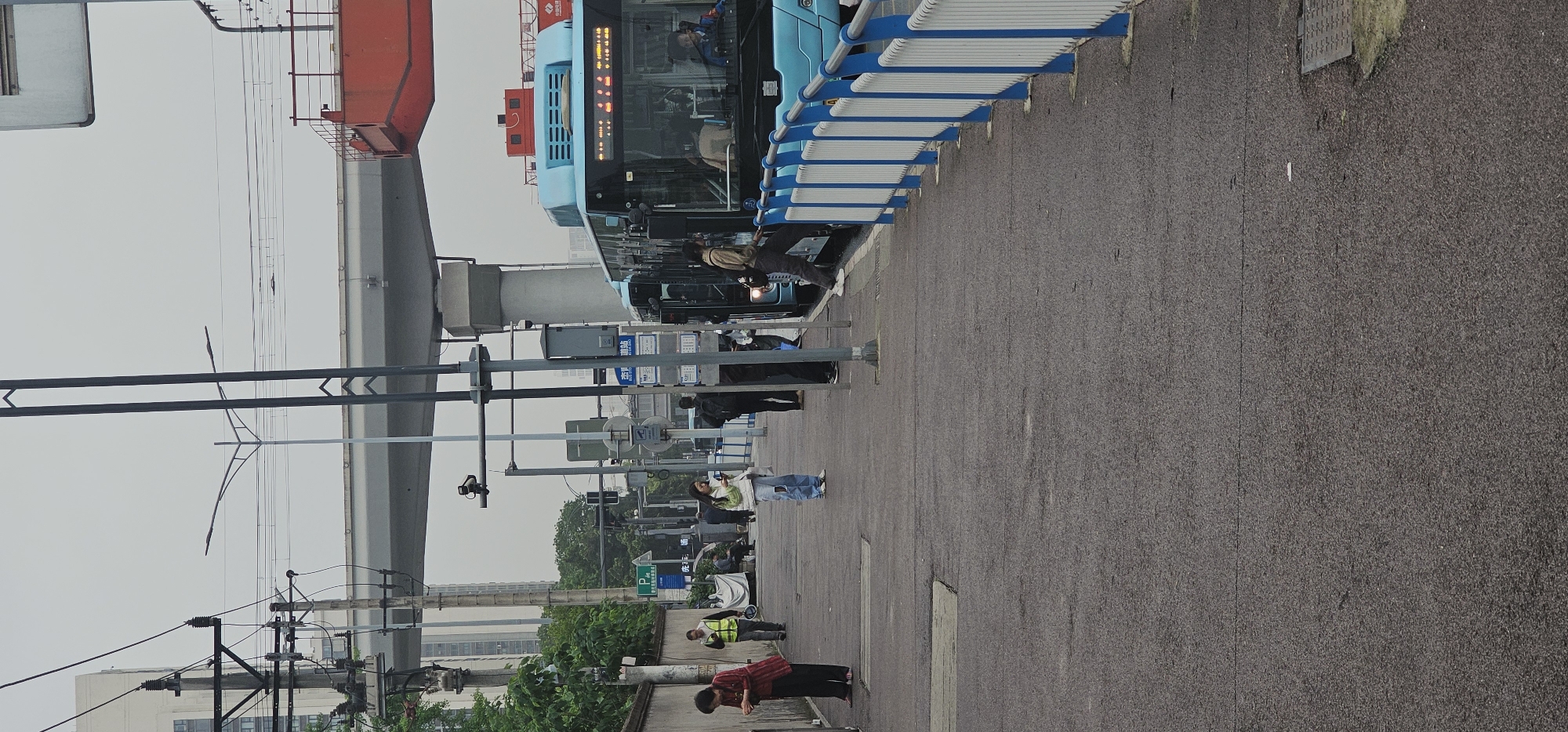 The image captures an urban street scene in Chongqing High-tech Zone, China, featuring a large, light blue public bus stopped at a bus station or stop. The bus, which has "H2" visible on its front, is positioned next to a long, blue and white striped shelter structure. The driver and some passengers are discernible inside the bus, and its destination sign displays illuminated Chinese characters.
Several individuals are present on the paved sidewalk. One person in a light brown jacket stands near the bus entrance, possibly in the process of boarding or alighting. Further along, a person wearing a white top and blue jeans is visible. In the foreground, a person in a red and black patterned shirt is bending down, while another individual in a yellow safety vest stands nearby.
Above the street, a prominent grey elevated concrete structure, potentially a monorail track or elevated roadway, spans across the scene. A large red, crane-like apparatus is visible atop this structure on the far left. The sky is overcast, suggesting daytime with diffused lighting. The area is equipped with multiple utility poles and overhead power lines. Various street signs with Chinese characters are present, including a green sign with a white 'P' indicating parking. The scene depicts a typical busy urban environment focused on public transportation.
