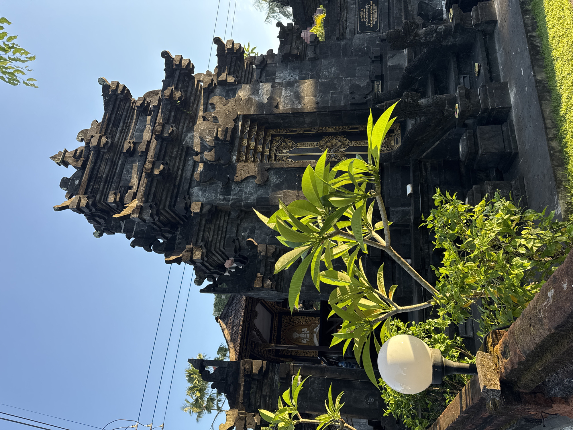 A street-level, outdoor photograph shows a tall, dark-colored stone structure that is likely a temple entrance or gate, situated in the city of Kuta, Indonesia. The structure is heavily decorated with layers of carved stone and intricate designs. A tall plumeria tree with bright green leaves is positioned to the right of the structure and a white, globe-shaped lamp is visible in the foreground. Part of the right side of the structure and a brick wall are covered in green foliage and grass. Power lines are visible against a clear blue sky in the upper left corner of the image. A smaller structure with a tiled roof can be seen further back through the temple gate. A black plaque with illegible white writing is affixed to the side of the structure.