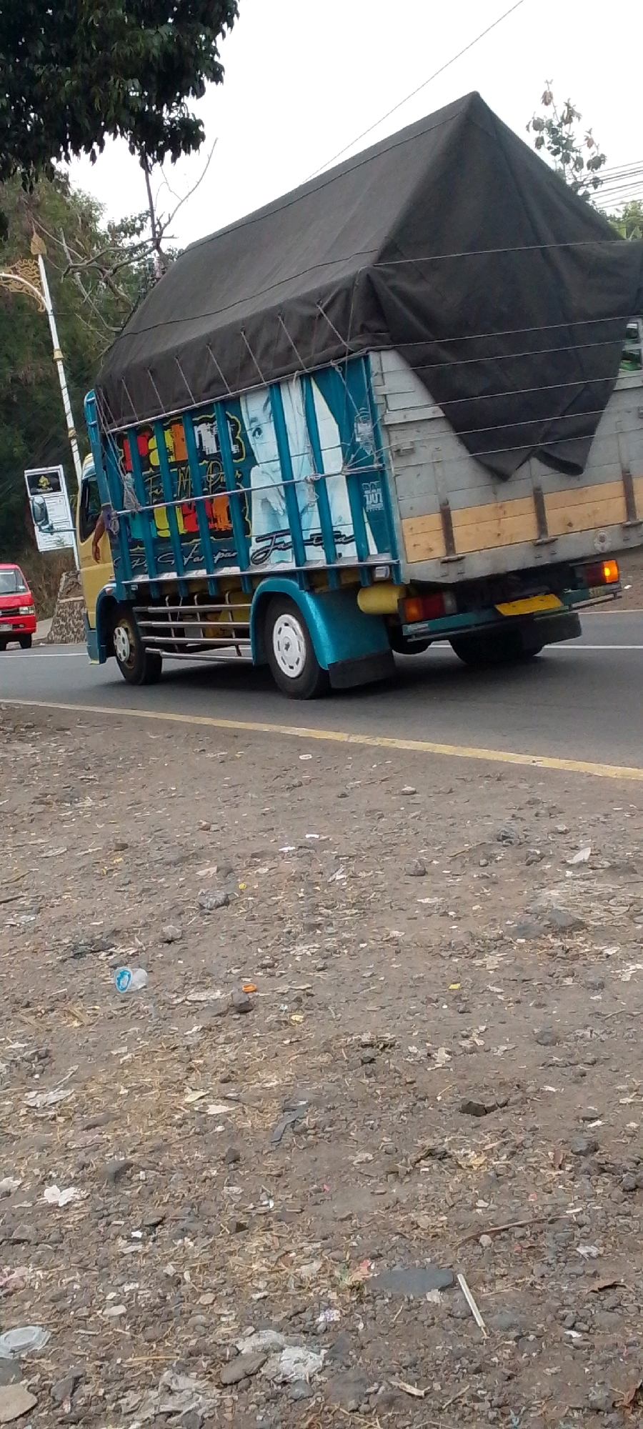 A truck is driving on a road in Cilaku, Indonesia. The truck is blue and yellow and has a black tarp covering its cargo. The truck is decorated with images of people and colorful patterns. In the foreground, there is a pile of dirt and debris. A red car is visible on the left side of the image.