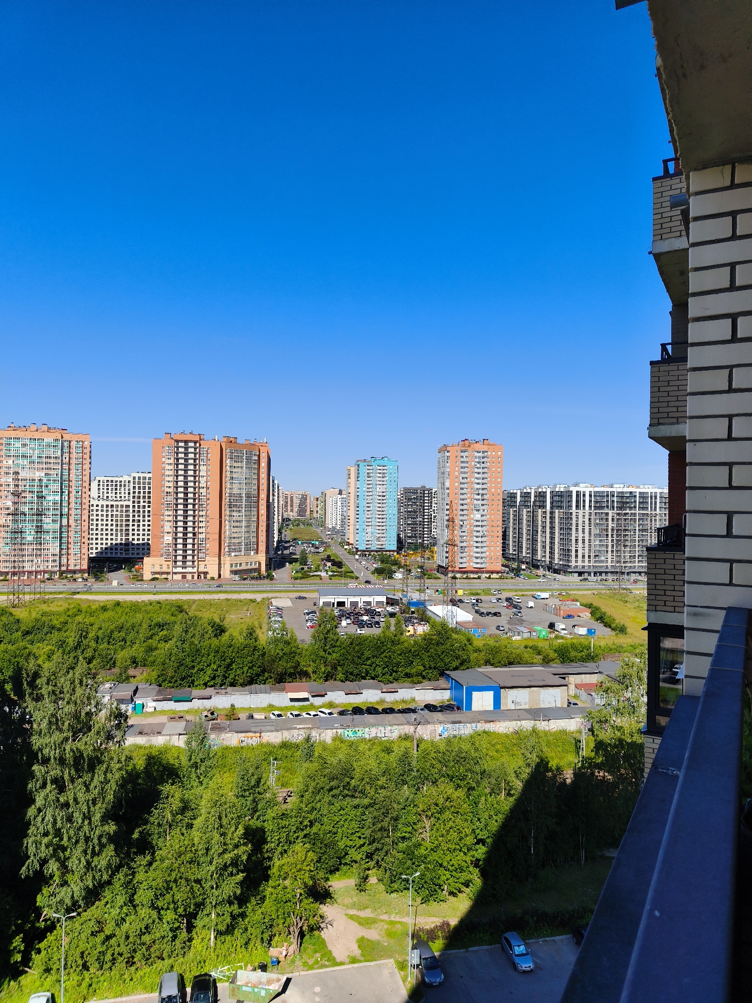 An aerial, full shot captures the cityscape of Murino, Russia, under a clear, bright blue sky. The foreground features the edge of a brick building under construction or renovation, with exposed concrete and metal railings. Beyond this, a vast expanse of lush green trees dominates the middle ground, interspersed with various buildings. Several multi-story residential buildings, characterized by their uniform modern architecture and diverse color palettes ranging from orange and white to blue and grey, are visible. A road stretches into the distance, with a noticeable presence of parked cars and some industrial-looking structures, including garages and warehouses, some adorned with graffiti. The overall scene depicts a developing urban area with a blend of residential and industrial elements.