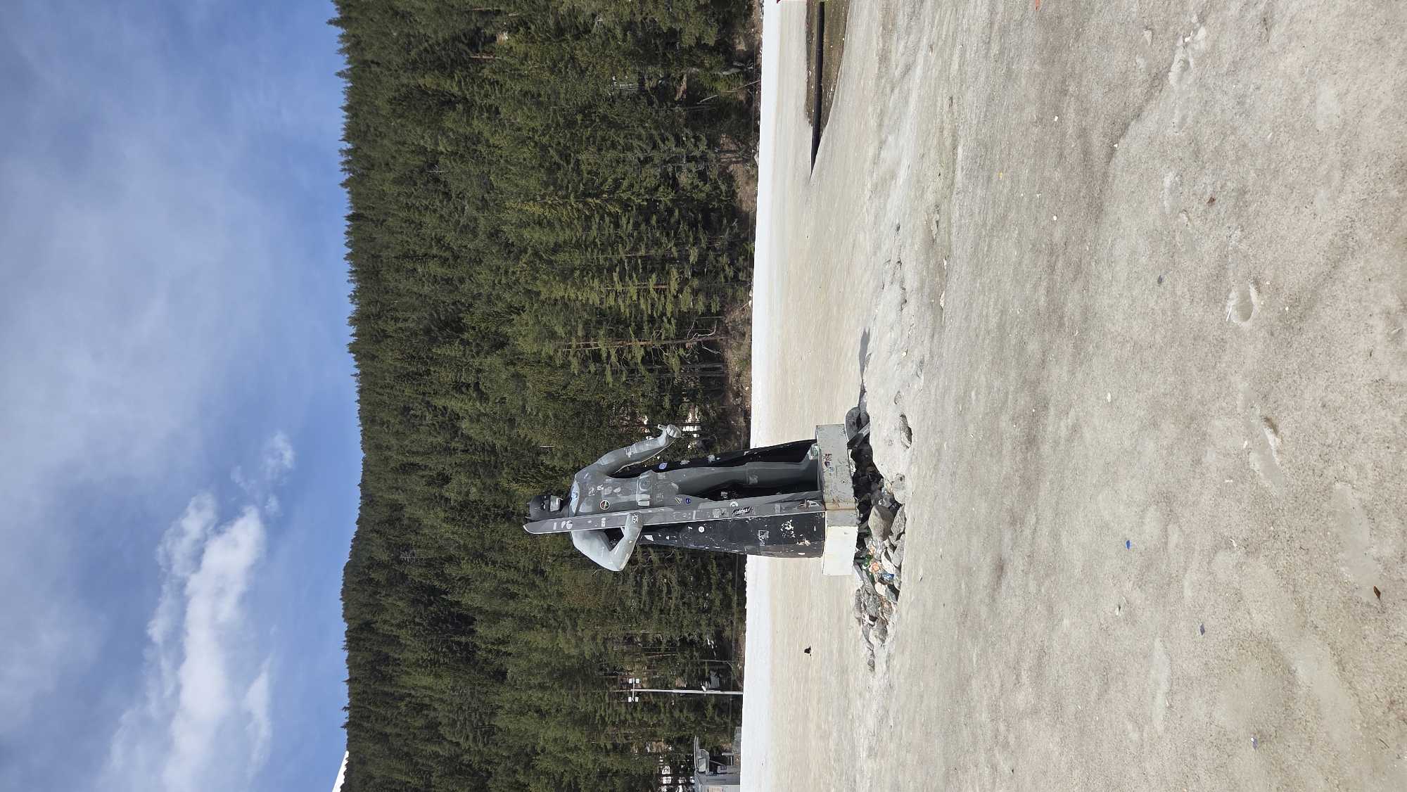 The image captures an outdoor scene dominated by a statue against a backdrop of a dense forest and a clear blue sky. The scene is oriented vertically, but should be viewed horizontally for correct interpretation.
In the foreground, a large statue depicts a person, likely an athlete, holding what appears to be a snowboard or skis. The statue is sculpted in a dynamic pose, suggesting movement. Its color is a dull metallic gray. The base of the statue is dark, possibly black, and is covered in numerous stickers and graffiti, indicating it's a popular or frequently visited spot. Around the base, there's a collection of debris, including what looks like plastic bottles and other refuse, suggesting litter has accumulated in a crevice of the ground.
The ground surrounding the statue is an expanse of dirty, grayish snow or ice, marked with footprints and uneven textures, suggesting it's either melting or heavily trafficked. In the midground, beyond the statue and the icy ground, a dense forest of coniferous trees stretches across the image, forming a dark green, solid line that contrasts with the brighter elements. The forest appears to cover a large hill or mountain.
Above the forest and the statue, the sky is a vibrant blue with sparse, wispy white clouds. The bright sky suggests it is daytime, likely during late morning or early afternoon, with clear weather conditions. The presence of snow/ice and the subject of the statue point to a winter or early spring setting in a mountainous or snowy region. No human activity is directly depicted, but the statue itself implies past or potential winter sports activity.
Visible text and symbols are present on the statue's base and equipment, mainly in the form of stickers. On the black pedestal, multiple stickers are visible, some circular, some rectangular. One sticker appears to have the letters "OG" and a symbol, while another shows a stylized mountain. On the snowboard held by the statue, there's a light-colored sticker, possibly a peace sign or a brand logo, though specific words are not clearly legible due to the distance and resolution.