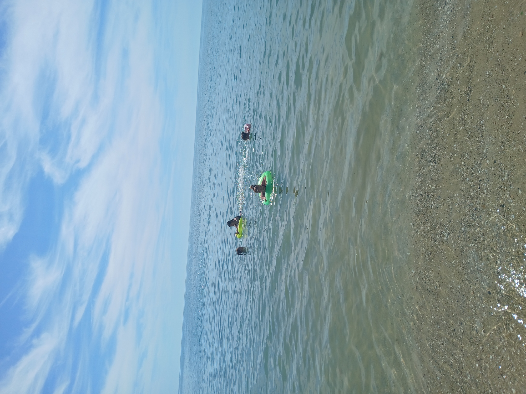 In Kranevo, Bulgaria, four individuals are observed in the calm, clear waters of the Black Sea. The scene is set under a bright, blue sky with scattered, wispy clouds. The shallow, sandy seabed is visible beneath the translucent water, with small pebbles and shells dotting the shore. Two individuals are swimming, while two others are using inflatable tubes. One person is in a green inflatable ring, another in a yellow one. The overall atmosphere is one of leisure and enjoyment in a coastal environment.