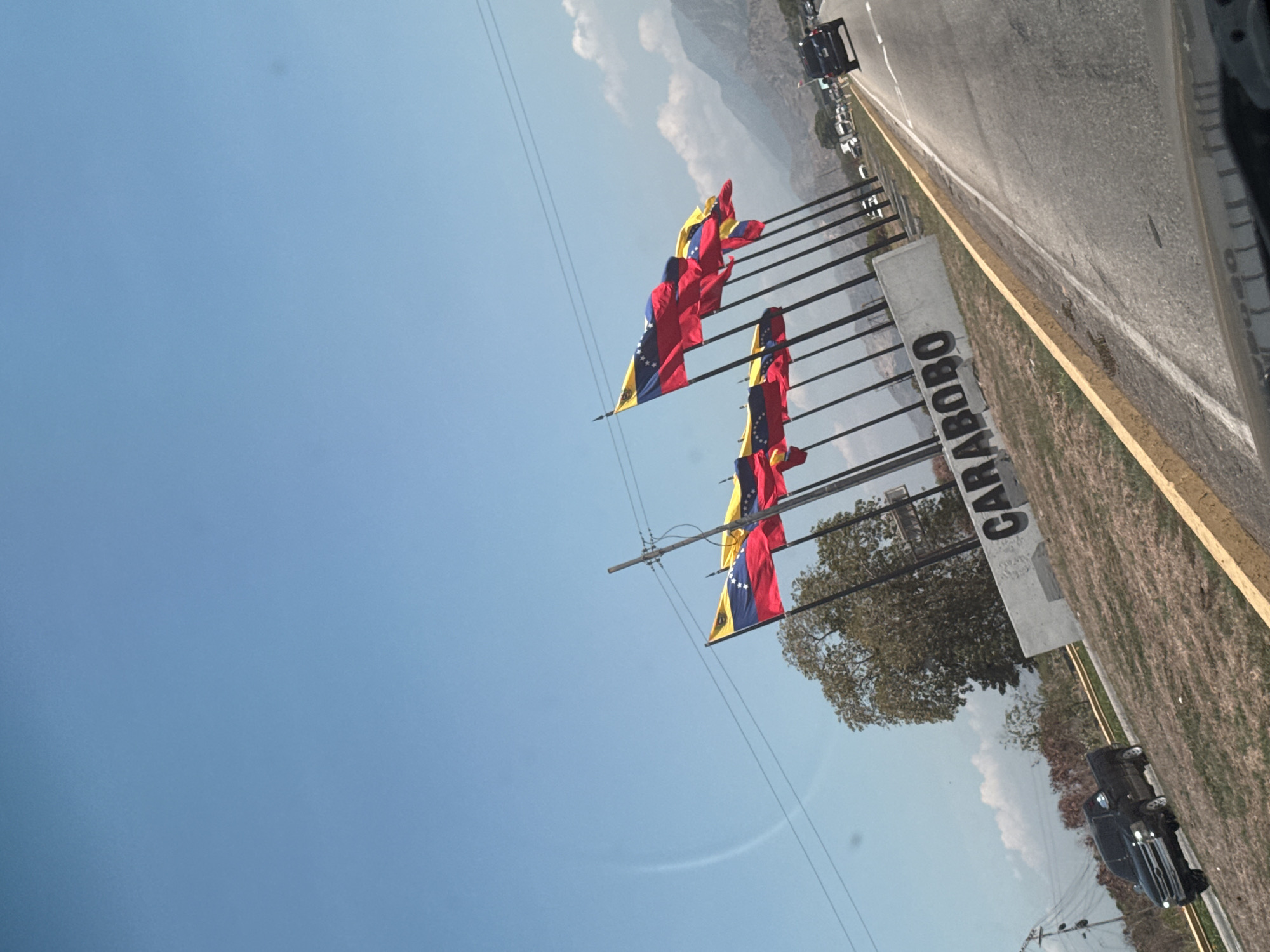The image shows an entrance to the city of La Cabrera, Venezuela. A series of Venezuelan flags wave in the wind next to a stone sign that says "CARABOBO". The photo appears to be taken from a moving vehicle along a highway with a yellow line marker. The background shows a mountainous terrain and a blue sky with scattered clouds. Some cars are visible on the road. The overall environment suggests a sunny day at a roadside location.