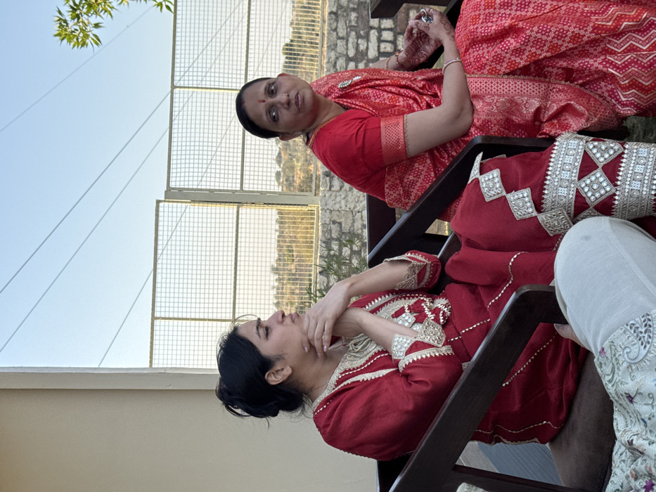 The scene depicts two women seated outdoors in Malasar, India. On the left, a woman with dark hair tied in a bun wears a deep red traditional Indian garment, embellished with intricate gold and silver embroidery and mirror work on the neckline, sleeves, and pants. She is seated in a dark brown wooden chair, with her hands clasped, and her gaze directed towards the right. Partially visible in the lower left foreground is the leg of a third individual, clad in light-colored patterned trousers. On the right, a second woman, identified by a red bindi on her forehead, wears a red and gold patterned saree over a red blouse. Her right hand, adorned with mehndi, holds a small silver object. She is seated in a dark brown wooden chair, looking forward. In the background, a wire mesh fence separates the seating area from an arid landscape featuring sparse dry vegetation and a stone wall, all beneath a clear blue sky traversed by utility lines. The area is illuminated by natural light.