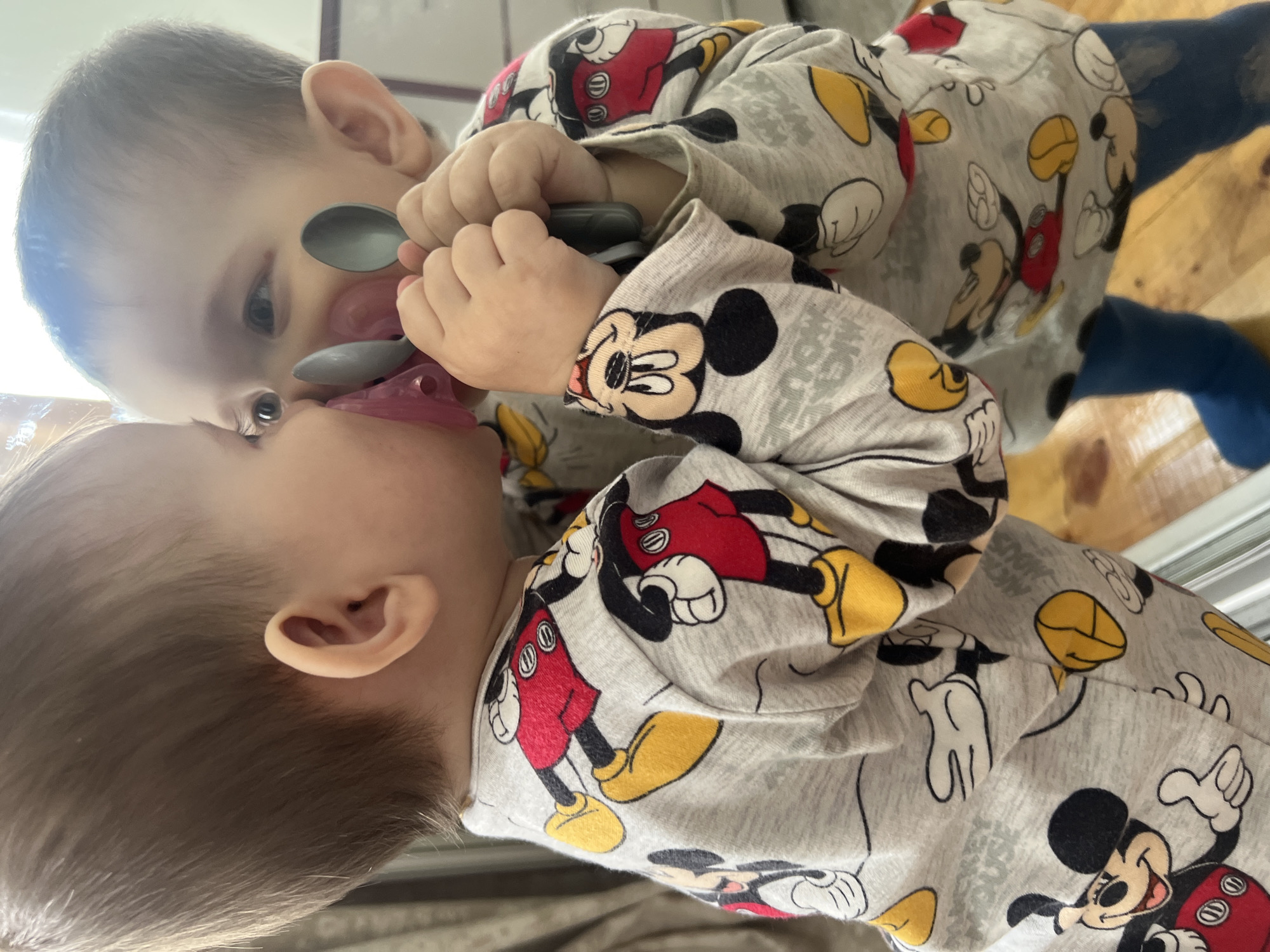 A toddler, wearing a gray long-sleeved shirt with a Mickey Mouse pattern and blue tights, is looking into a mirror. The toddler is holding two spoons and has a pink pacifier in their mouth. The toddler's reflection is visible in the mirror, which appears to be leaning against a wall. The background is blurred but suggests an indoor setting with some curtains and furniture. The toddler's hair is brown, and they have light-colored eyes. The lighting in the image is natural, coming from the right side, casting soft shadows.