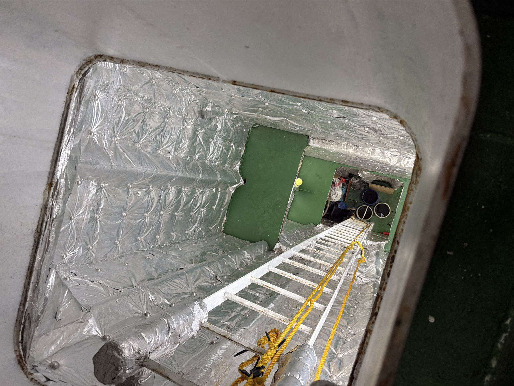 This image depicts a vertical shaft within a structure, likely on a vessel or industrial building. A white metal ladder with yellow ropes attached extends down into the shaft. The walls of the shaft are lined with quilted silver insulation material. At the bottom of the shaft, a green-painted floor is visible, with some objects scattered on it, including dark containers and what appears to be some equipment. The overall impression is one of an internal, utilitarian space. The location in Melaya, Indonesia, suggests this could be part of a port facility, a fishing vessel, or an industrial site within the town. There are no people visible in the image.