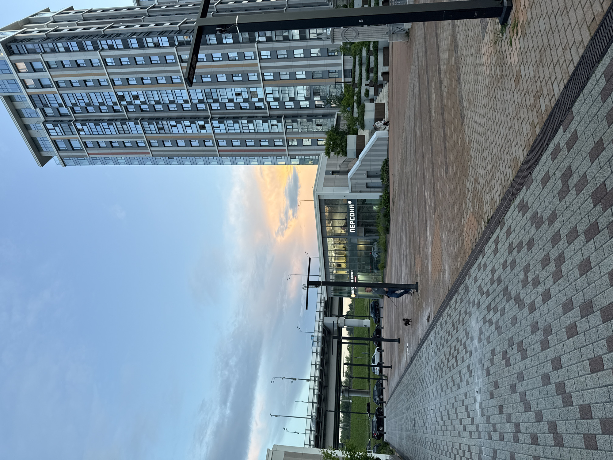 The image depicts an outdoor urban scene in Moscow, Russia, featuring a modern high-rise apartment building and a low-rise commercial building with glass facades. The sky shows a mixture of blue hues and diffused sunset colors with clouds. A pedestrian walkway paved with interlocking bricks in a geometric pattern leads into the scene. A man is walking a small black dog on a leash on this walkway. In the background, a multi-lane overpass with streetlights spans across the image. Cars are parked and driving on a road beneath the overpass. The commercial building has signage that appears to read "ПЕРСОНА" (Persona) and "ЦВЕТОМАРКЕТ" (Flower Market). The architectural style is contemporary, with clean lines and reflective surfaces.