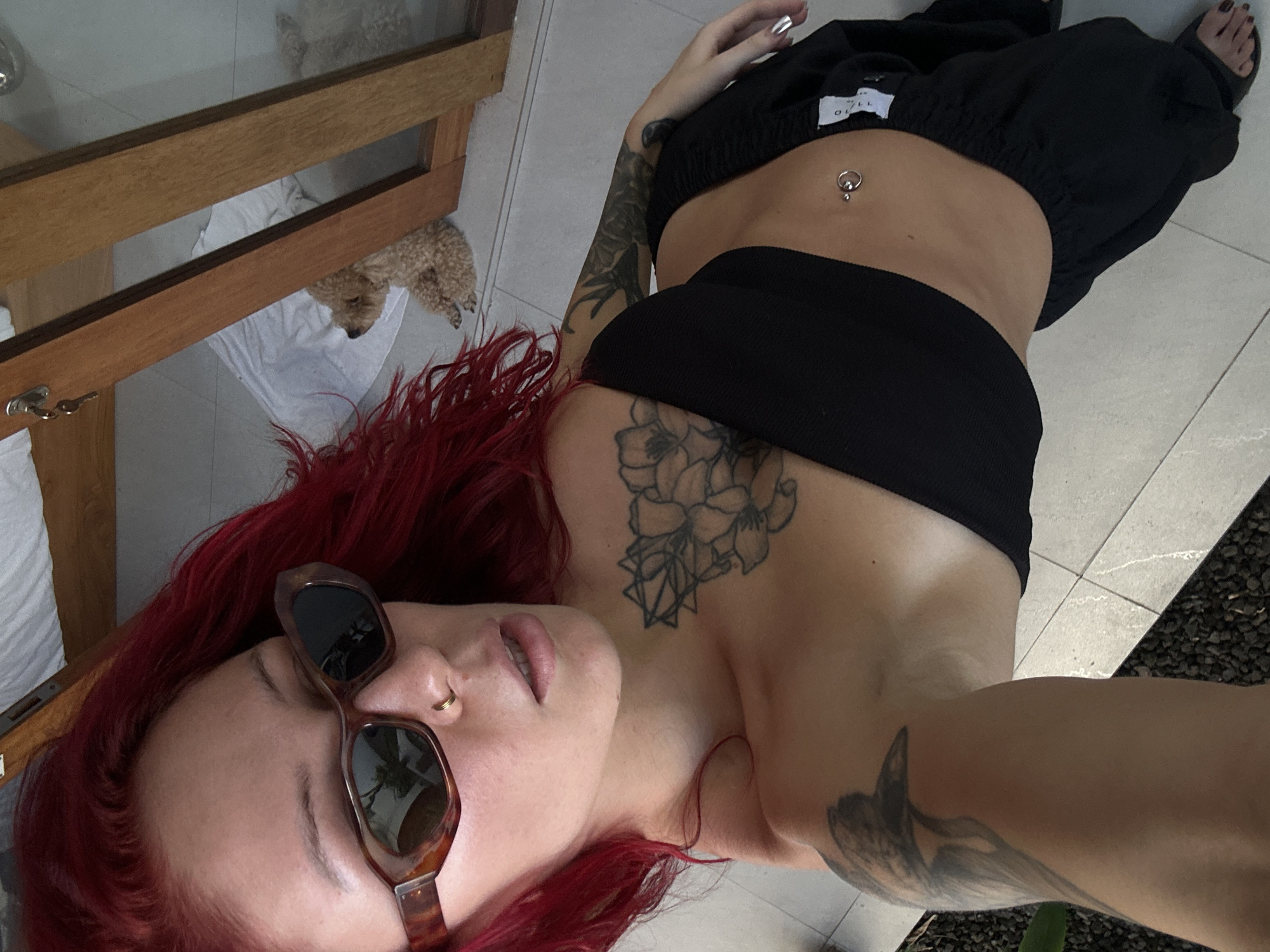 A person with long, bright red hair, wearing brown tortoiseshell sunglasses, takes a selfie while lying down or reclining. She has a small gold hoop nose piercing and a silver navel piercing. Her attire consists of a black bandeau top and black loose-fitting pants with a visible white label that reads "ORLL" or similar text. Several tattoos are visible: a floral and geometric design on her left upper chest/shoulder, and extensive sleeve tattoos on both arms, with an indistinct creature on the left arm.
The setting appears to be an indoor or semi-outdoor space with light-colored tiled flooring. In the background, a small, light brown curly-haired dog is resting on a white cloth, partially visible through a wooden frame, possibly part of a door or large window. The wooden frame includes a lock with a key. To the person's right, an area with dark gravel or stones and some sparse greenery suggests an adjacent outdoor space. The natural light indicates it is daytime. The overall mood is relaxed and casual. The location context is North Kuta, Indonesia.