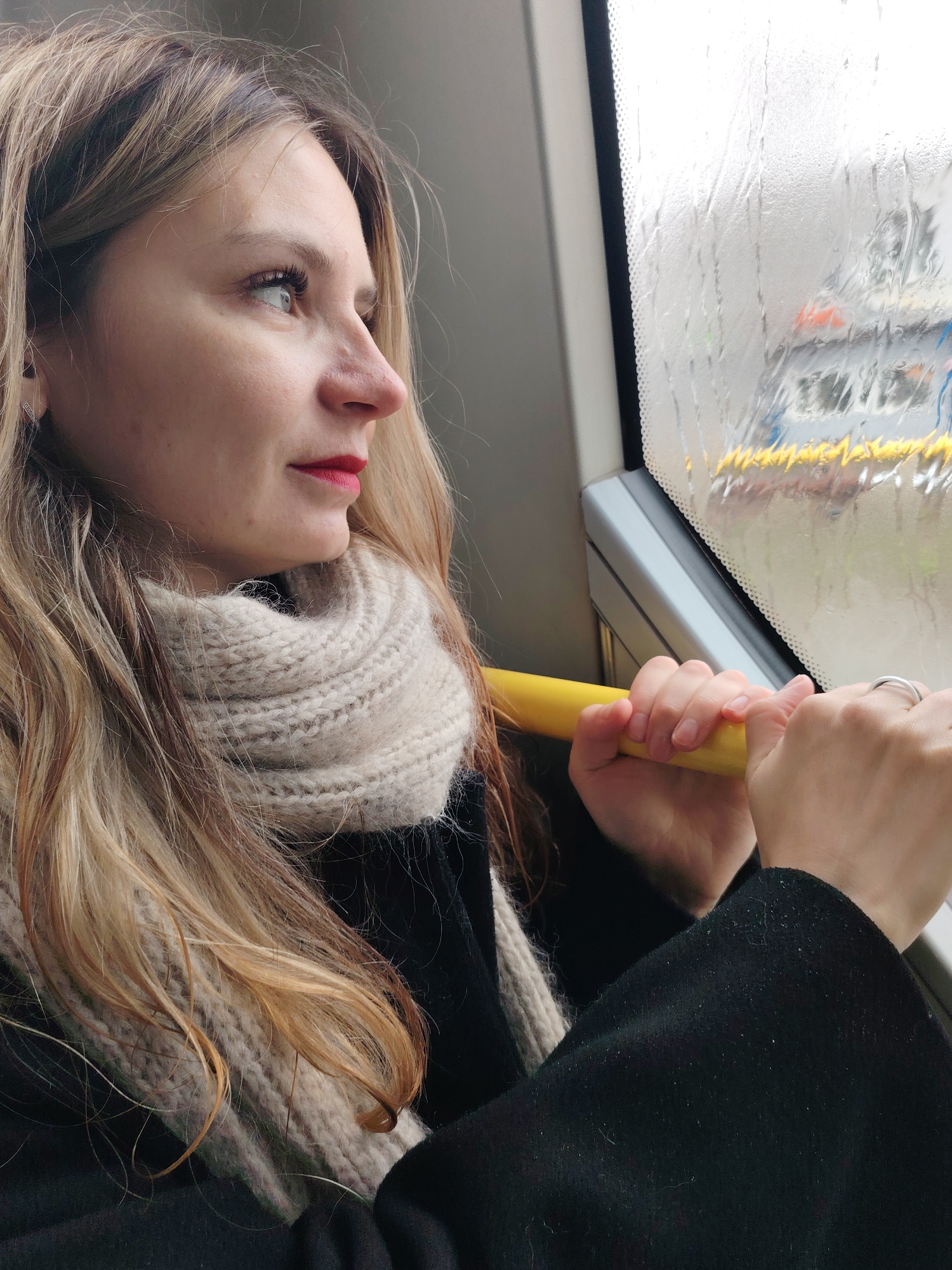 A young woman with long blonde hair is looking out of a rain-streaked window. She is wearing a black coat and a light-colored, chunky knit scarf. She is holding onto a yellow pole, possibly on a bus or train. Outside the window, a blurry view of a vehicle with yellow accents can be seen. The location is identified as the city of Chaikovsky, Russia.