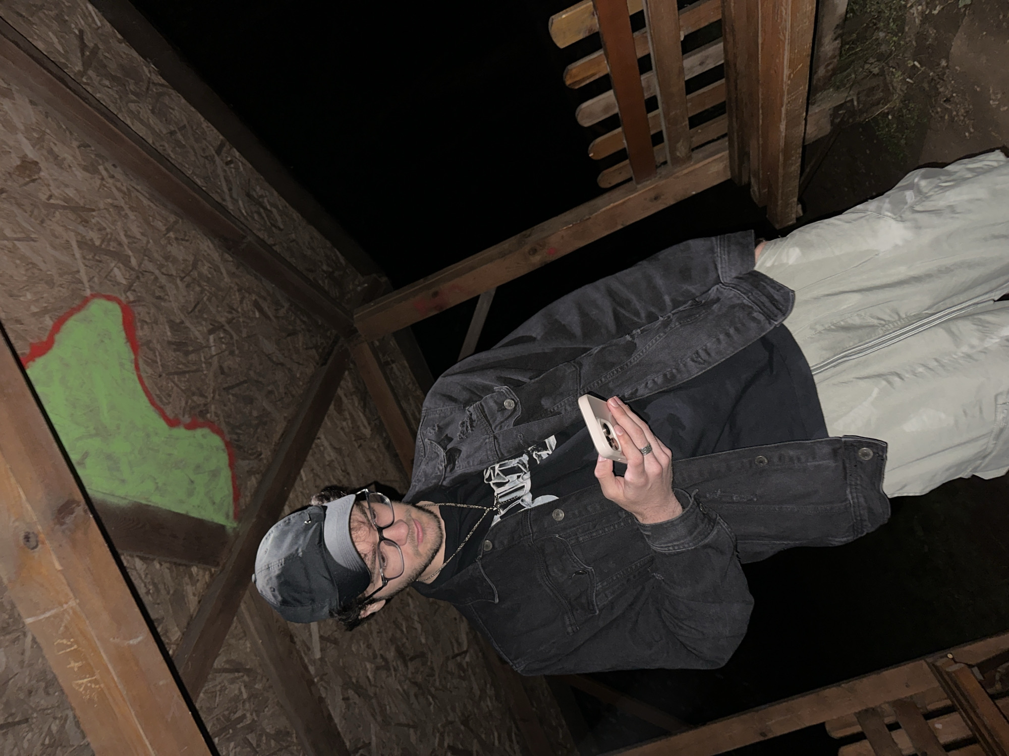 The image features a young man captured at night within a rustic wooden structure, likely an outdoor shelter or gazebo. He is wearing a dark cap, glasses, a black t-shirt with an indistinct white graphic, a distressed black denim jacket, and light-colored pants. A chain necklace is visible around his neck, and a silver ring adorns his right ring finger. He is holding a light-colored smartphone with a clear case in his right hand, looking intently at its screen.
The setting appears to be an outdoor area at night, with the background beyond the wooden structure completely dark. The structure itself is composed of oriented strand board (OSB) or similar plywood walls and robust wooden beams. On the OSB wall to the left, there is a prominent piece of graffiti: a large, irregular green shape outlined in red. Faint carvings, including what looks like "J+K" followed by a heart, are visible on a wooden beam near the man's head. Parts of a wooden fence or railing are also visible to the right. The man's expression is neutral and focused on his device. The location is Blagoevgrad, Bulgaria.