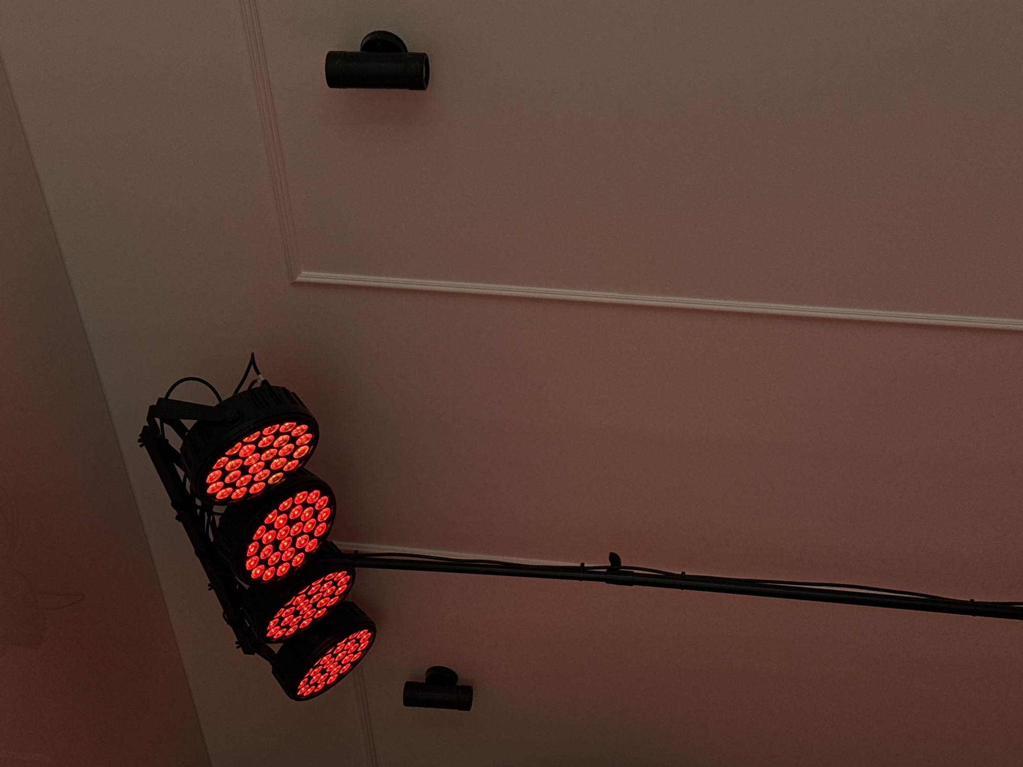 The image shows a stage lighting setup with four red LED par lights mounted on a bar. The lights are angled upwards and are illuminating a pale pink wall with white molding. A black pole supporting the lighting bar extends downwards, with cables visible. On the wall to the right, a dark cylindrical light fixture is mounted. To the left, another dark cylindrical light fixture is mounted. The lighting and wall suggest an indoor environment, possibly a studio or event space. There are no people or animals present. The image is a close-up, medium shot, capturing the lighting equipment and a portion of the wall. The location is not specified in the image or the provided context.