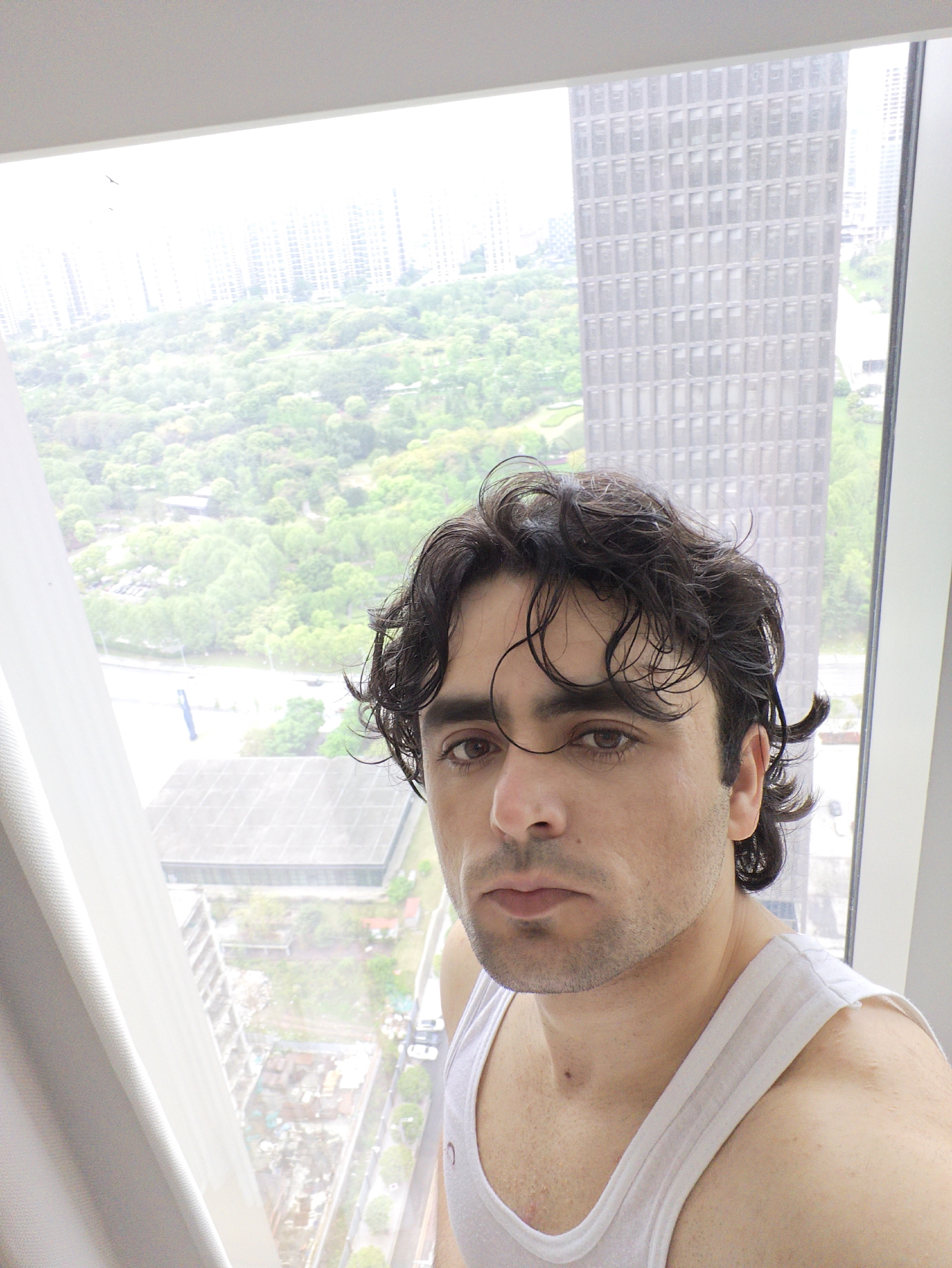 A medium-shot selfie of a Caucasian man with dark wavy hair, dark eyes, and light stubble, wearing a white tank top. He is standing in front of a large window with white framing, presumably indoors. Through the window, a cityscape is visible. A tall, rectangular skyscraper with numerous small, square windows is prominent on the right side of the view. In the background, additional buildings are visible, along with a large area of green vegetation, possibly a park or wooded area. A highway or large road can be seen winding through the trees. The light suggests it is daytime, but the overall tone is overcast or slightly hazy. Based on the context provided, the location is likely in the Liangjiang area of China.