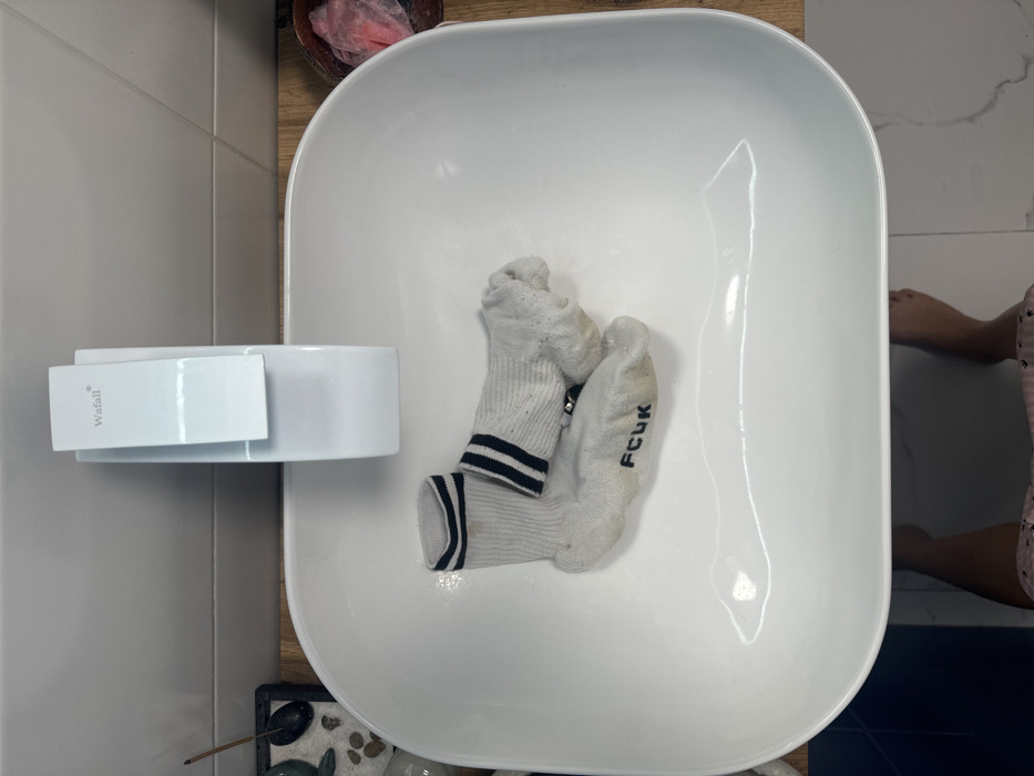 A pair of worn white socks with black stripes and the letters "FCUK" are placed inside a white, rounded rectangular sink. The sink has a white, minimalist faucet attached to its side. The surrounding environment includes a wooden countertop, a tile wall, and a small decorative arrangement of stones and a stick. A glimpse of a person's lower legs can be seen on the right edge of the image.