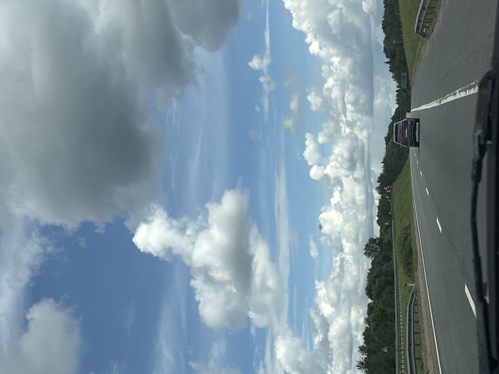 The image shows a highway with a dark SUV driving away from the camera. The road has white lane markings and guardrails. On either side of the road are green trees and grass. The sky is blue with scattered white and gray clouds. The image appears to have been taken from the perspective of another vehicle. There are no visible people in the image. The location is identified as Leshkovo, Russia.